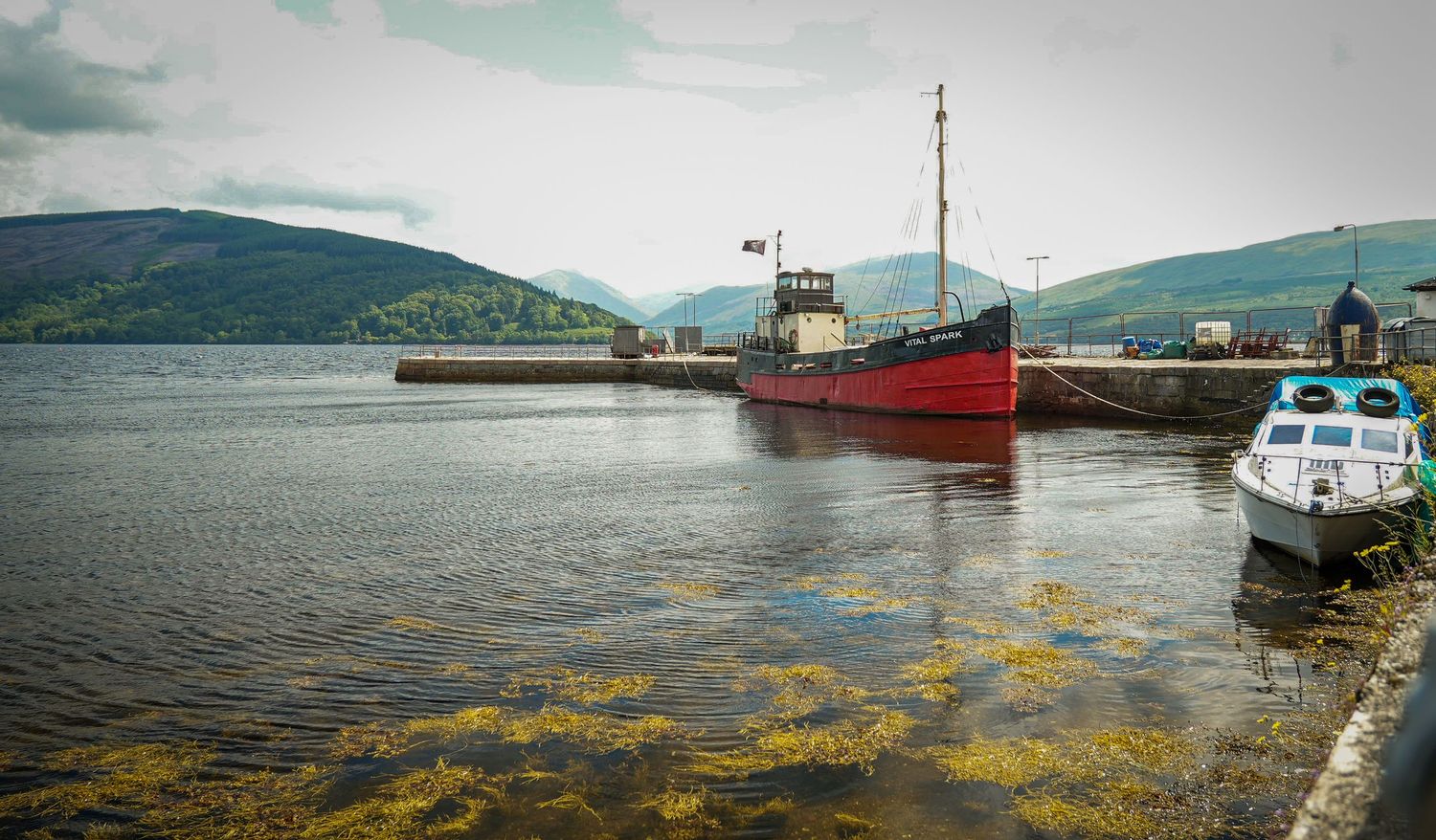 Red fishing vessel moored at a quiet Scottish harbor with misty hills and seaweed floating in the calm water.