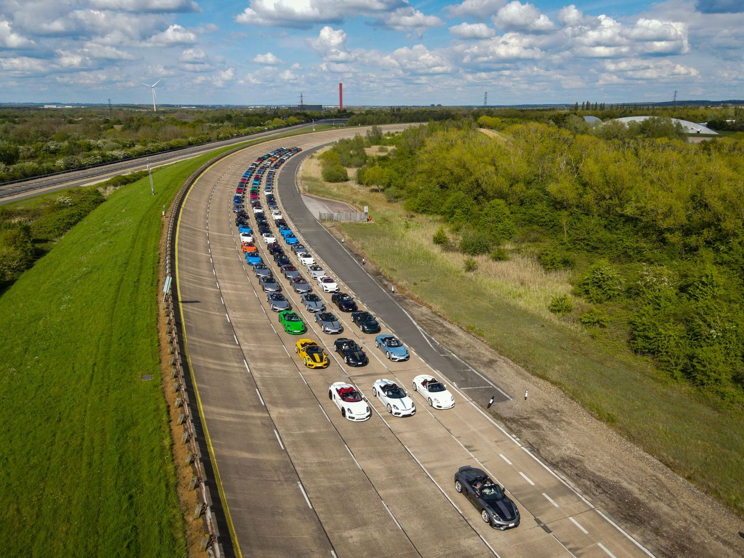 Aerial view of cars lined up on a highway with green surroundings and puffy clouds in a blue sky.