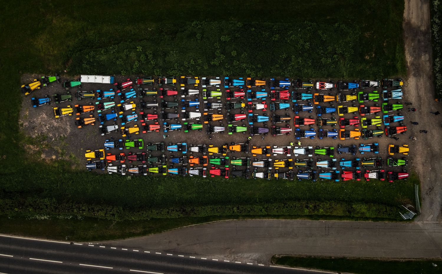 Aerial view of colorful model cars arranged in neat rows on grassy ground alongside a road.