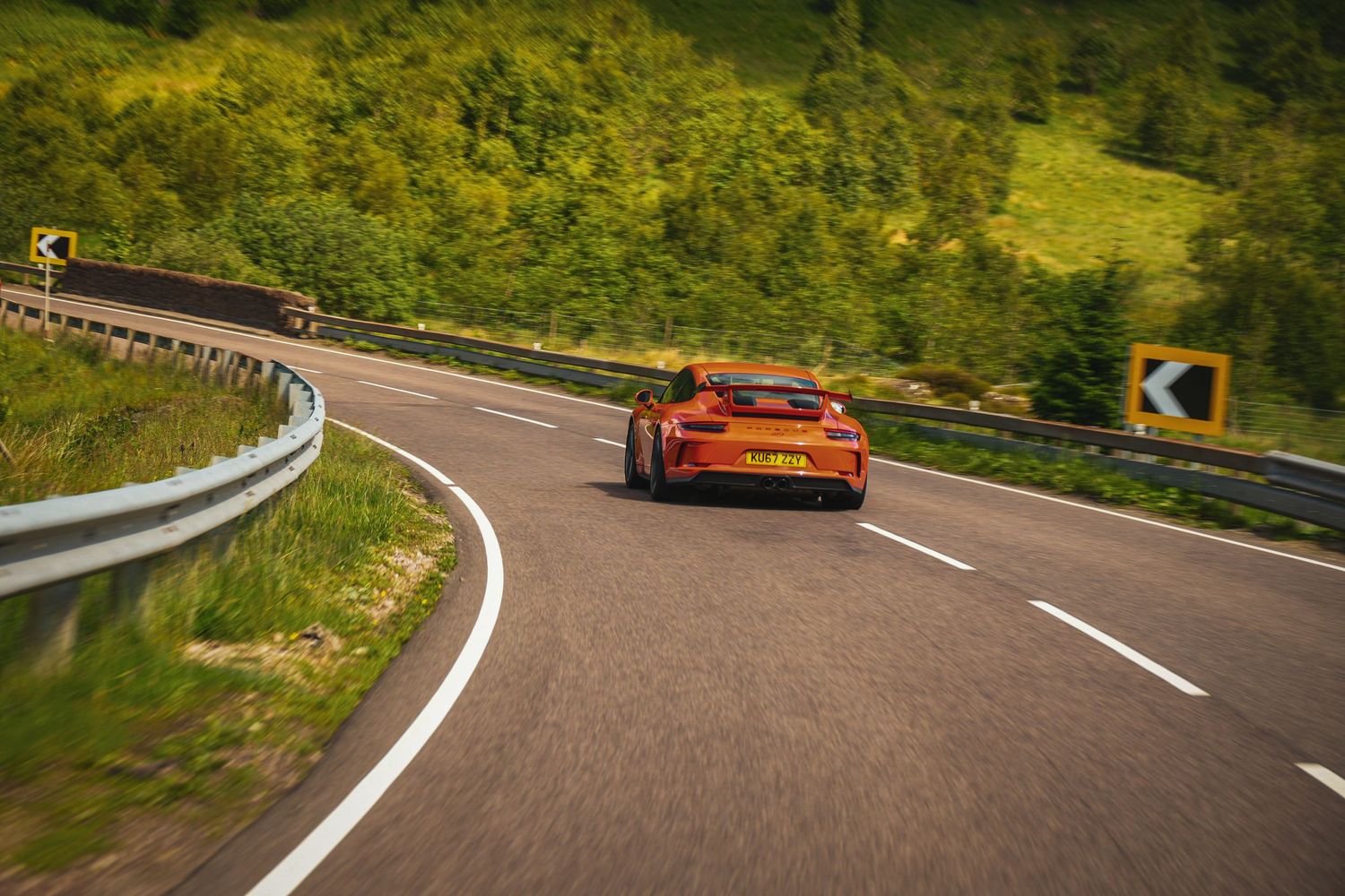 An orange sports car drives along a winding mountain road with lush green scenery and guardrails.