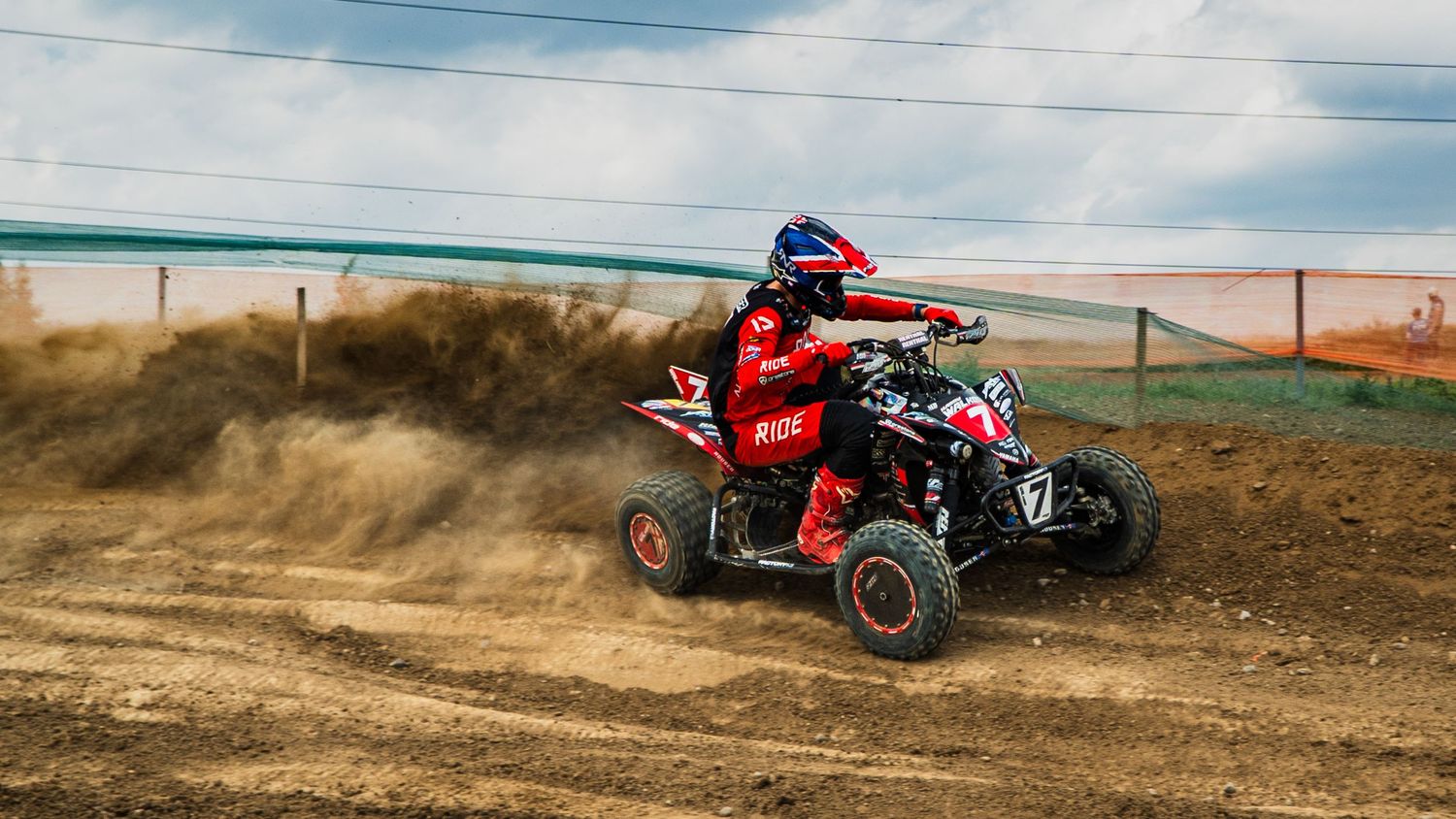 A rider races across a dirt track on a red ATV quad bike kicking up dust clouds in an action-packed moment.