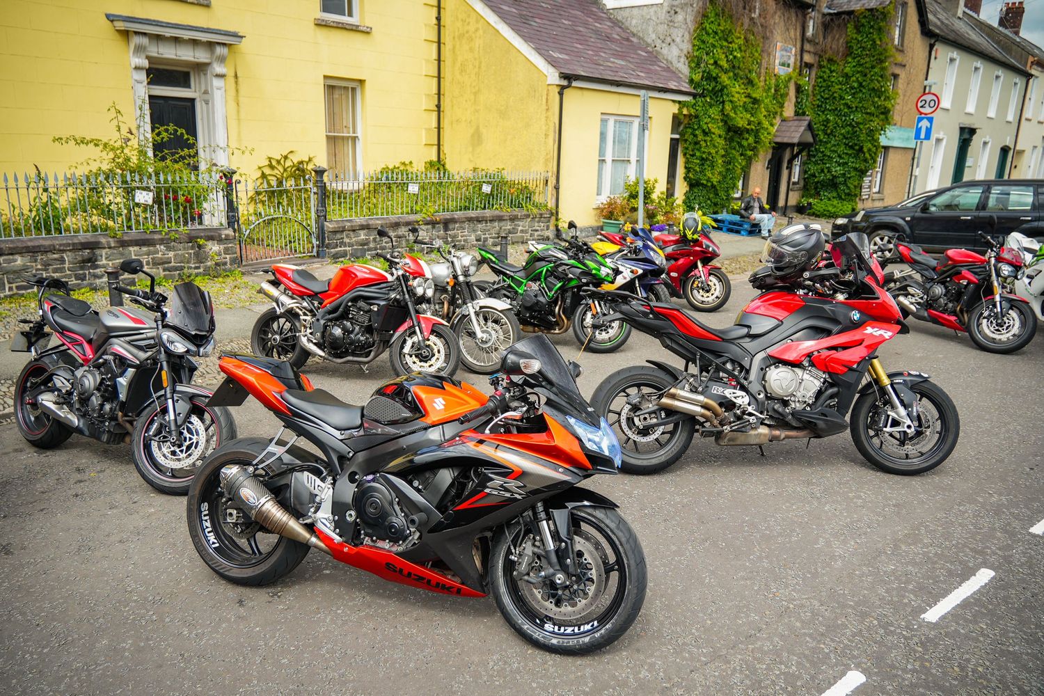 A row of colorful motorcycles parked along a street with yellow buildings and parked cars in the background.