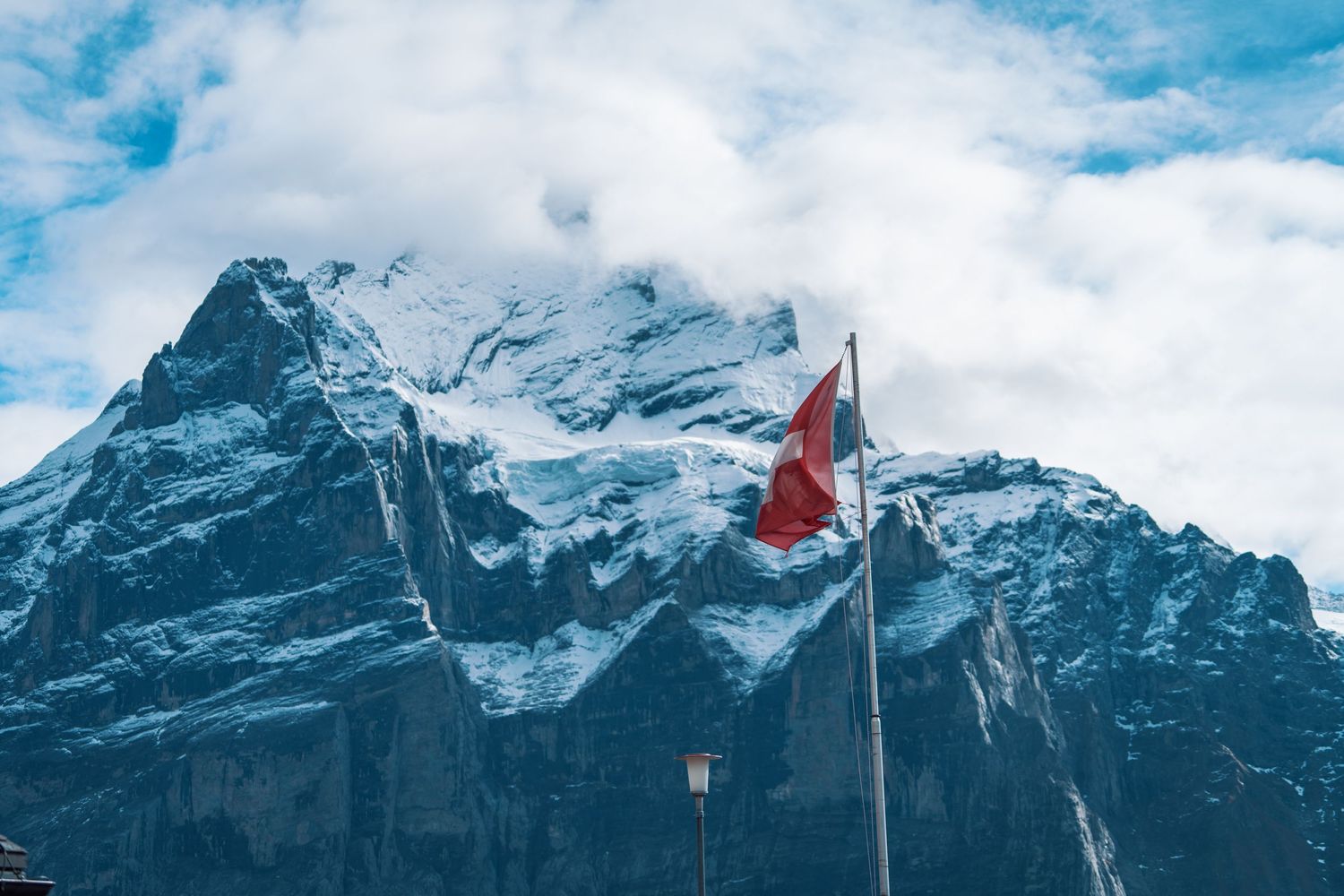 A Swiss flag waves against a backdrop of snowy mountain peaks and cloudy skies in the Swiss Alps.