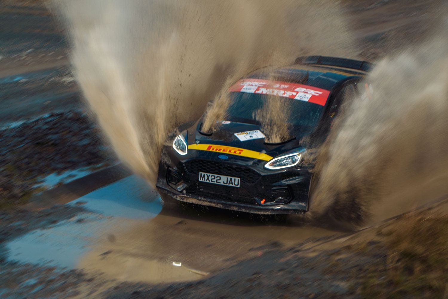 Rally car kicks up dramatic clouds of dirt and water while racing through muddy conditions on a course.
