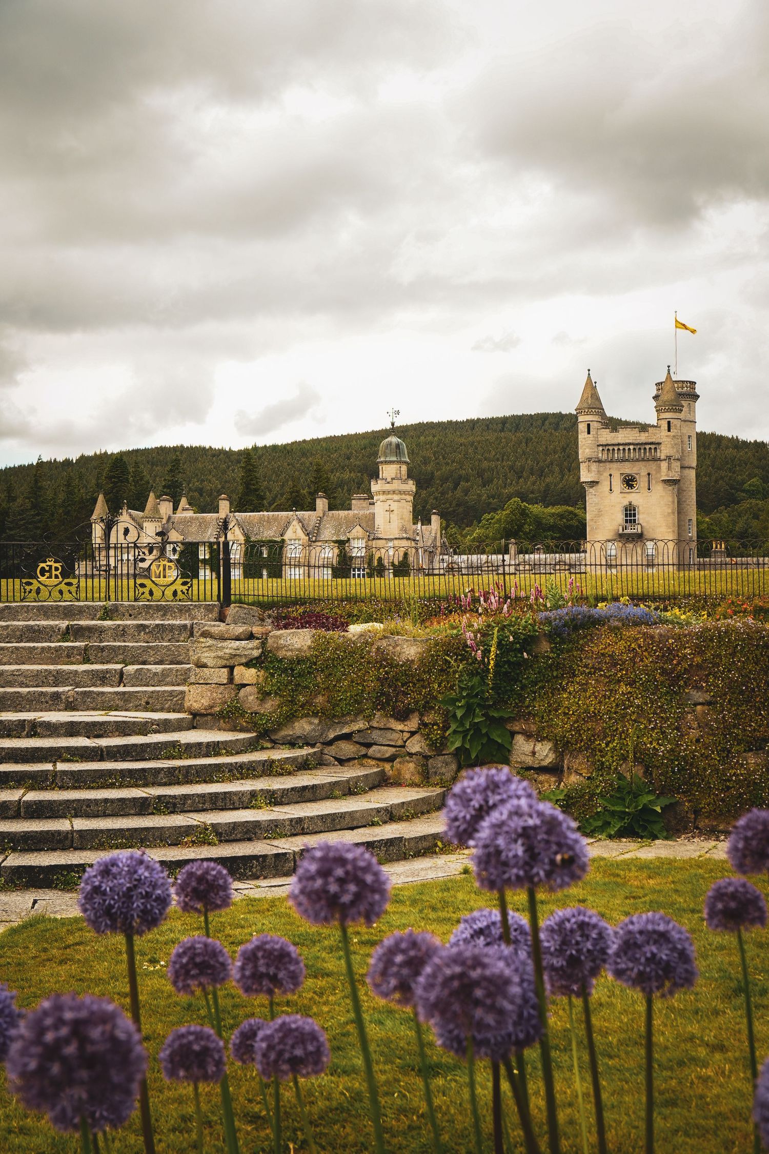 Balmoral Castle in Scotland with purple allium flowers in foreground and stone steps against moody cloudy sky.
