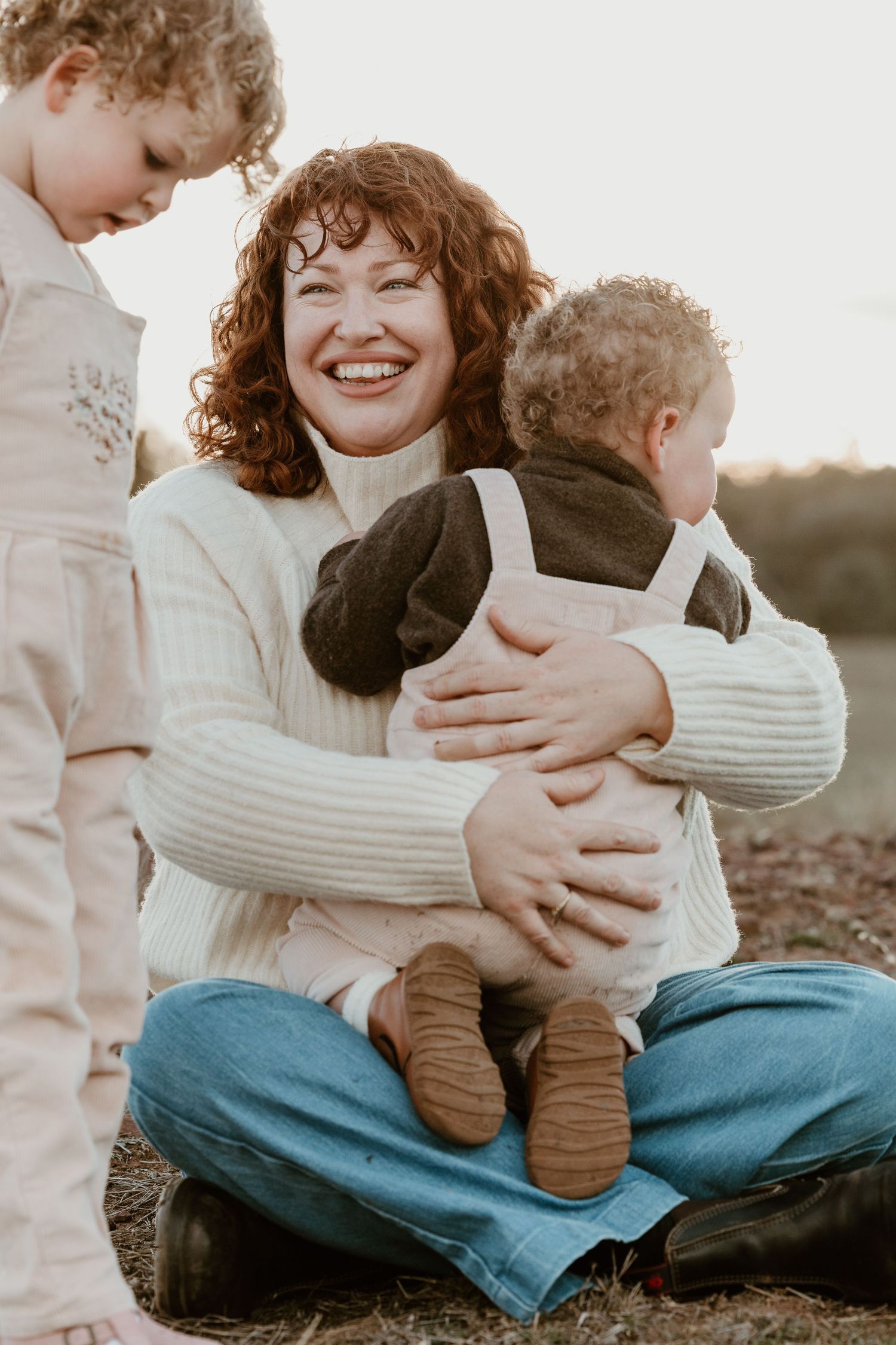 Smiling redhead woman sitting cross-legged outdoors, hugging a baby in overalls while a toddler stands nearby.