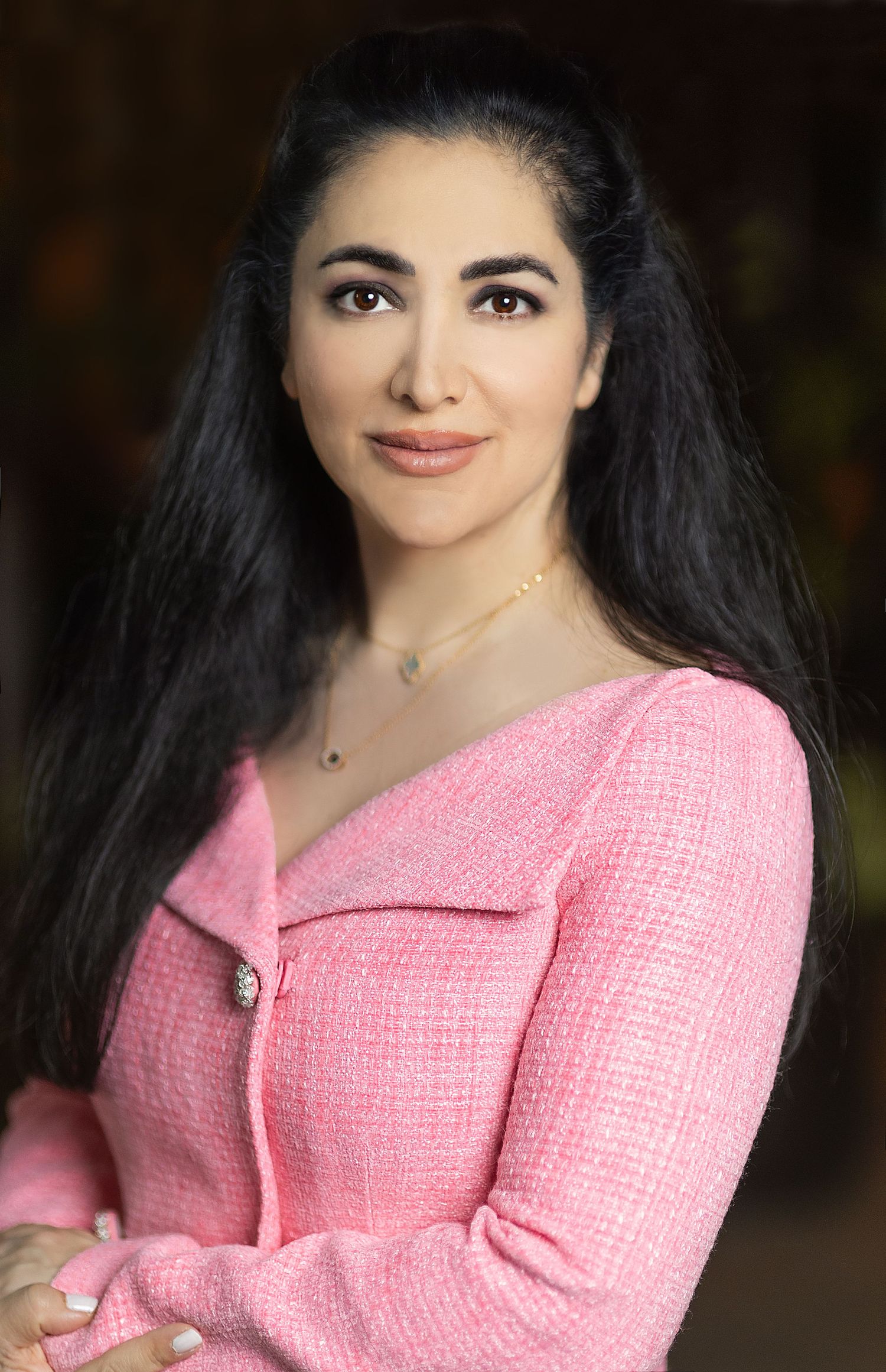Portrait of woman with long dark hair wearing a pink tweed jacket against blurred background.