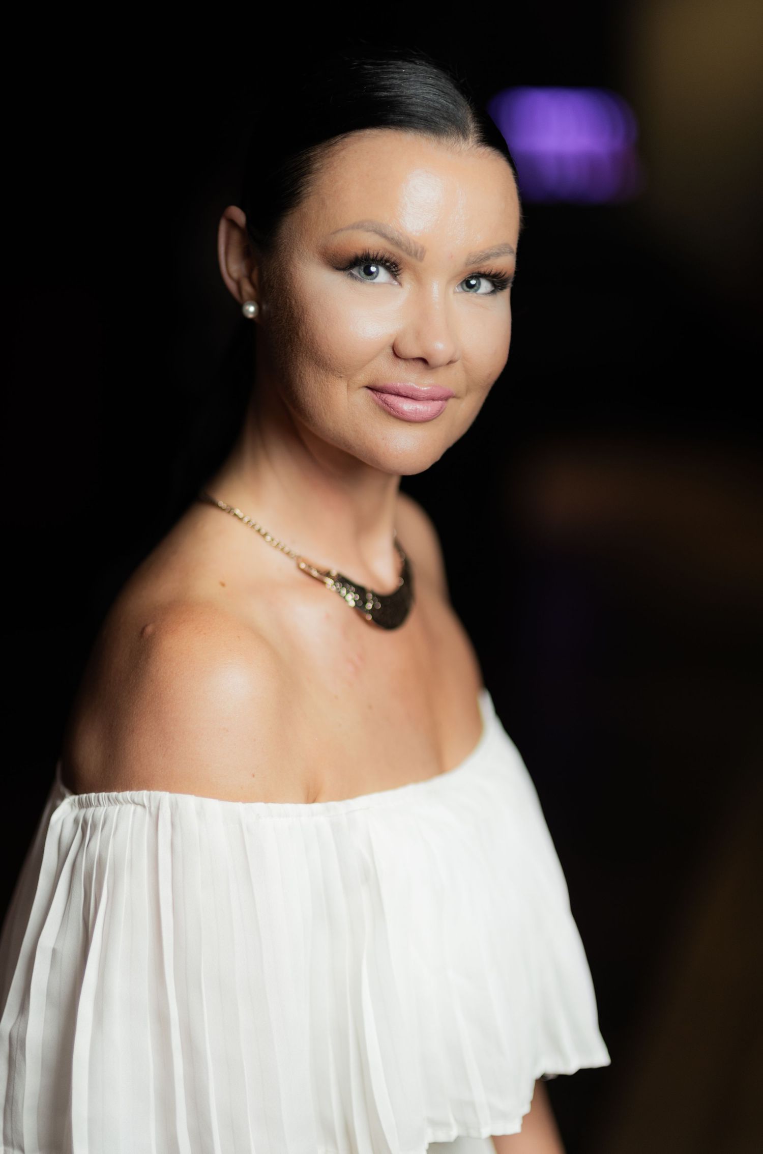Portrait of woman in white off-shoulder top wearing a sparkling necklace against dark background.