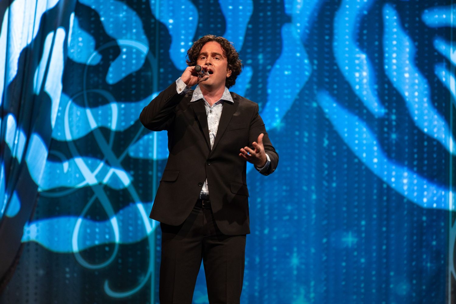 A performer in a black suit gestures while singing on stage with a vibrant blue backdrop display.