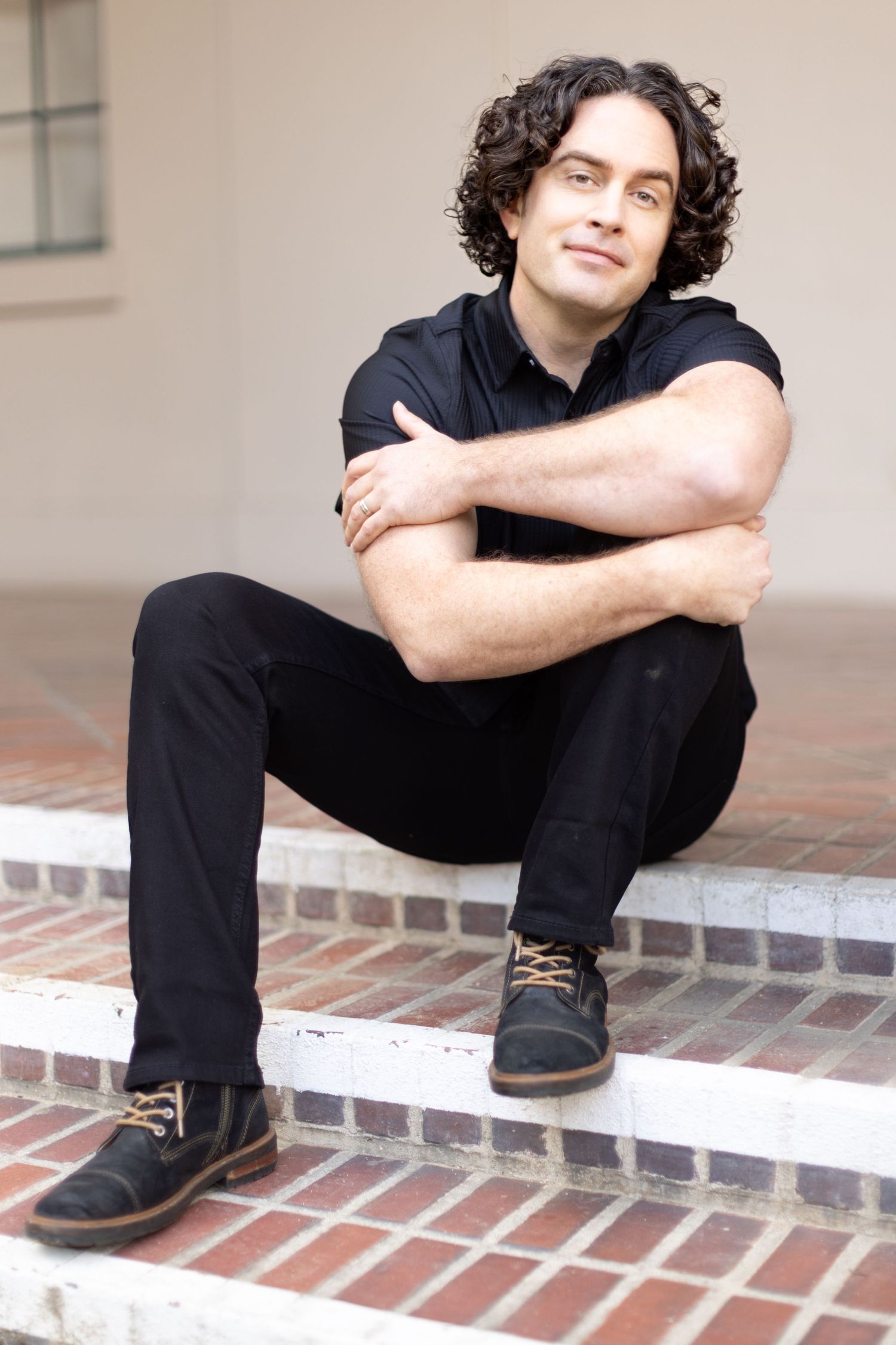 A person in all black attire sits casually on brick steps with arms crossed, wearing dark casual shoes.