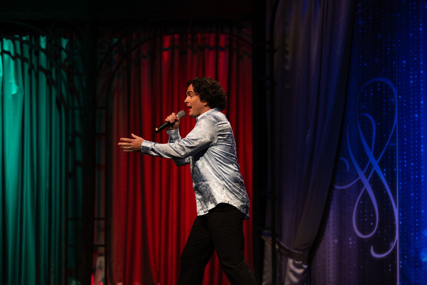A performer gestures energetically on stage with colorful red, green and blue curtains in the background.