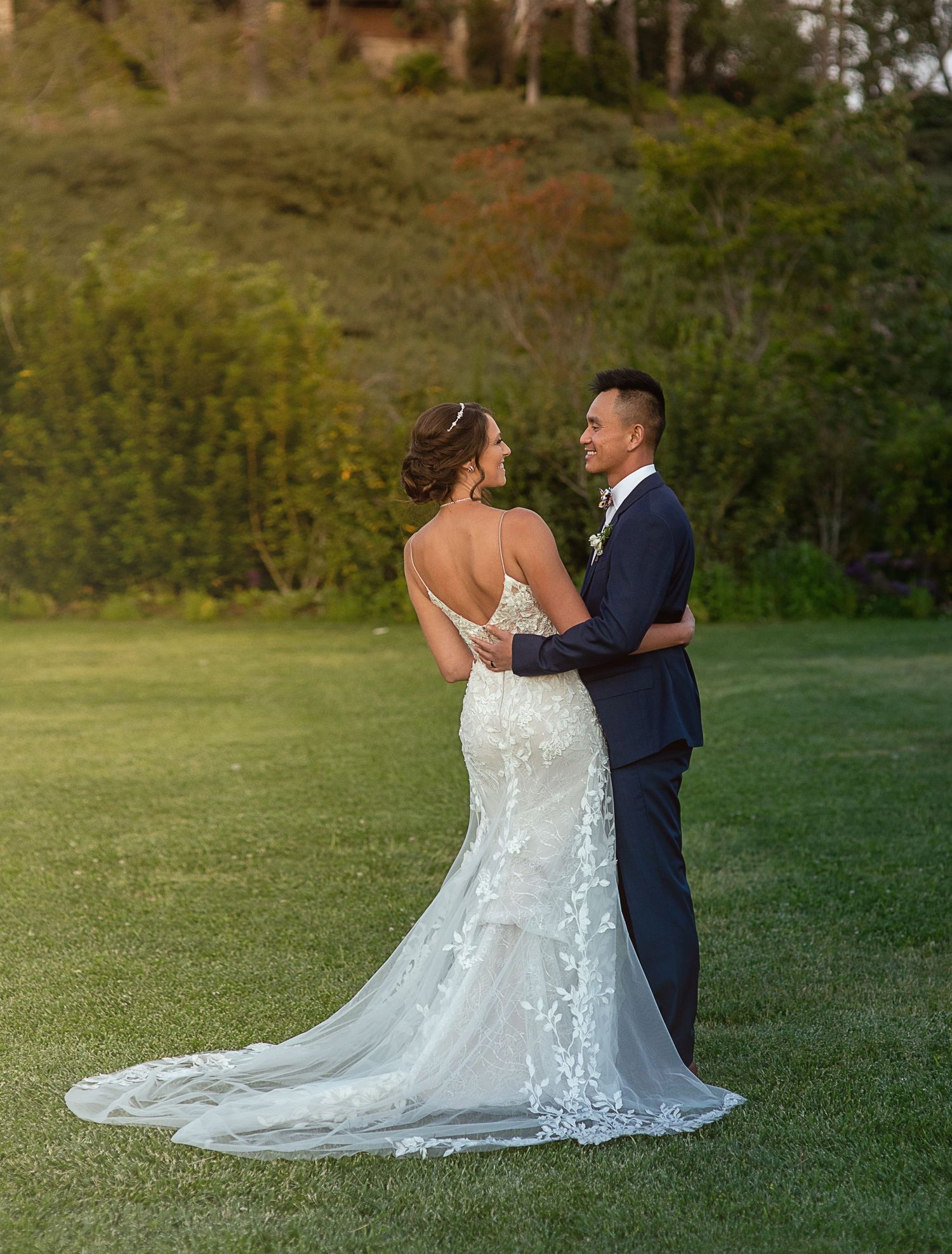 A romantic wedding photo on a grassy lawn showing the back of a lace wedding dress with a long train.