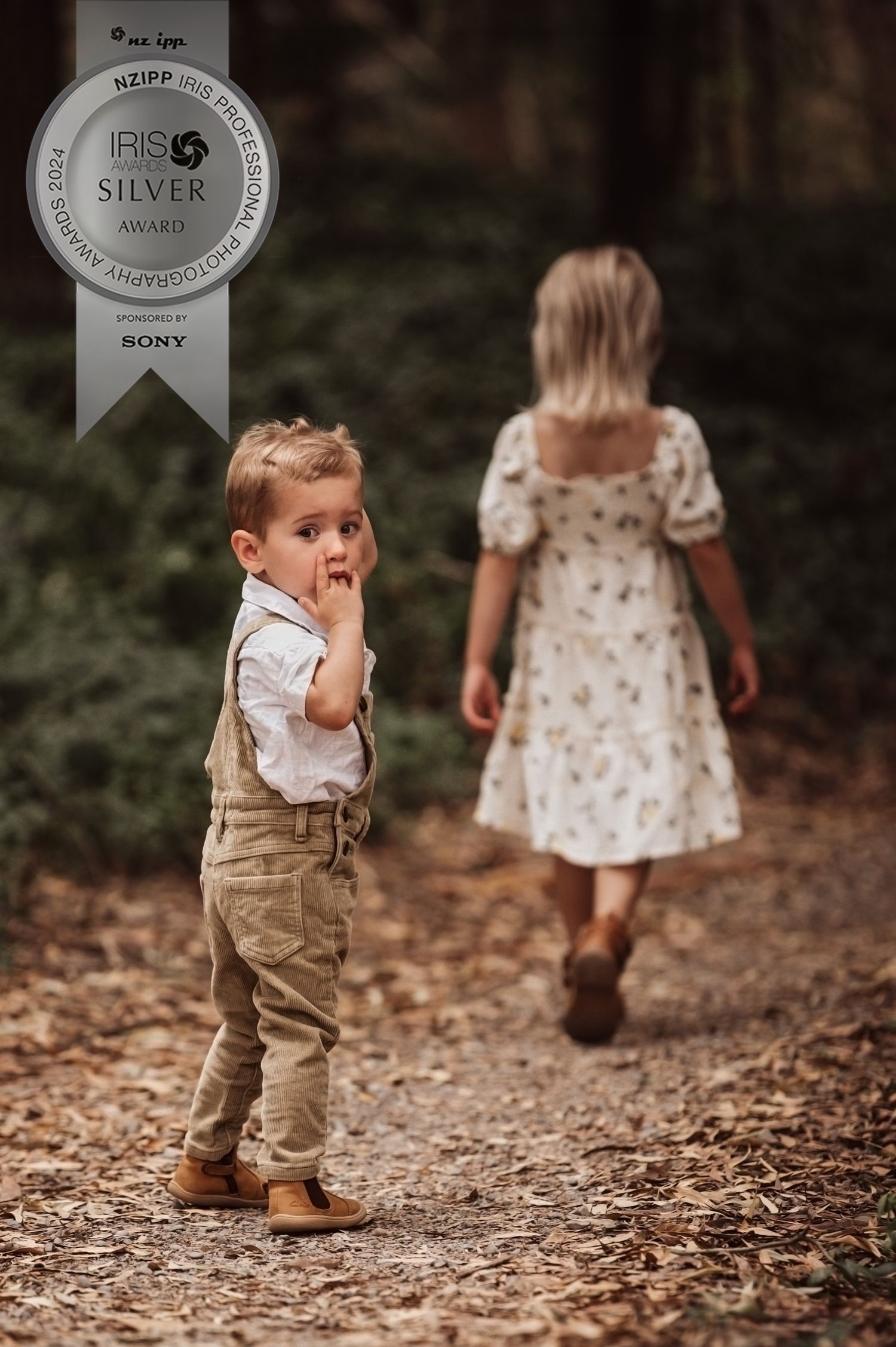 Young children walk along a leaf-covered forest path, with one wearing khaki overalls and another in a floral dress.