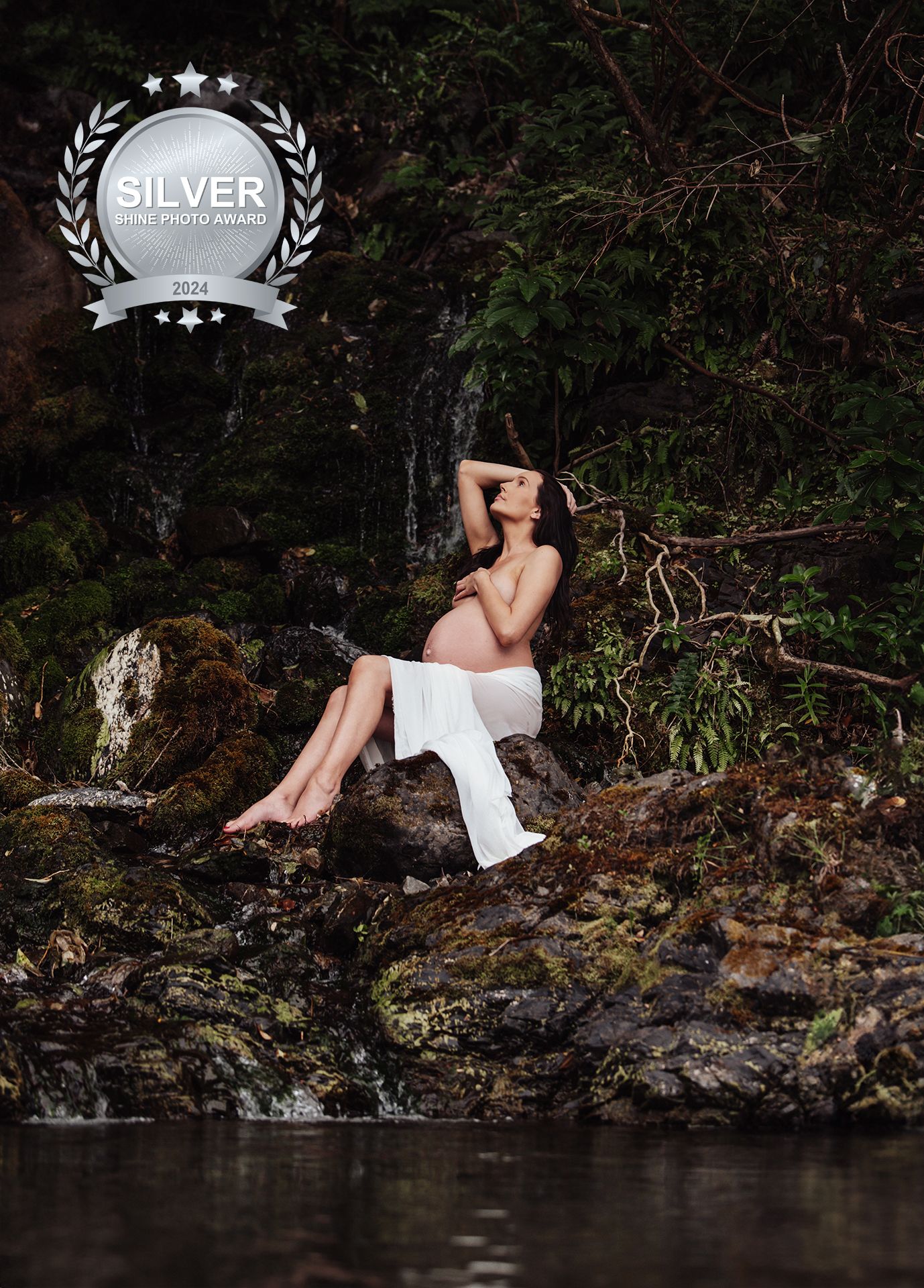 Artistic maternity photo of woman in white dress sitting by a serene waterfall.