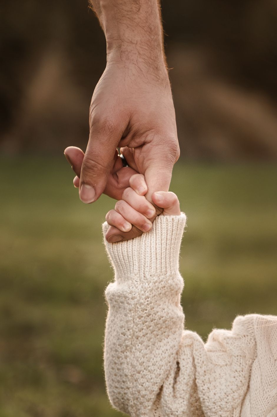 Close-up of an adult hand holding a small hand wearing a cream knit sweater outdoors in soft lighting.