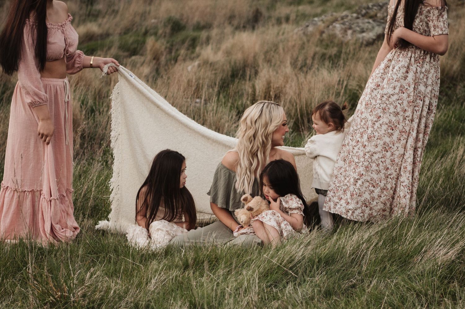 A group relaxes on a white blanket in a grassy meadow while wearing flowing floral dresses in muted tones.