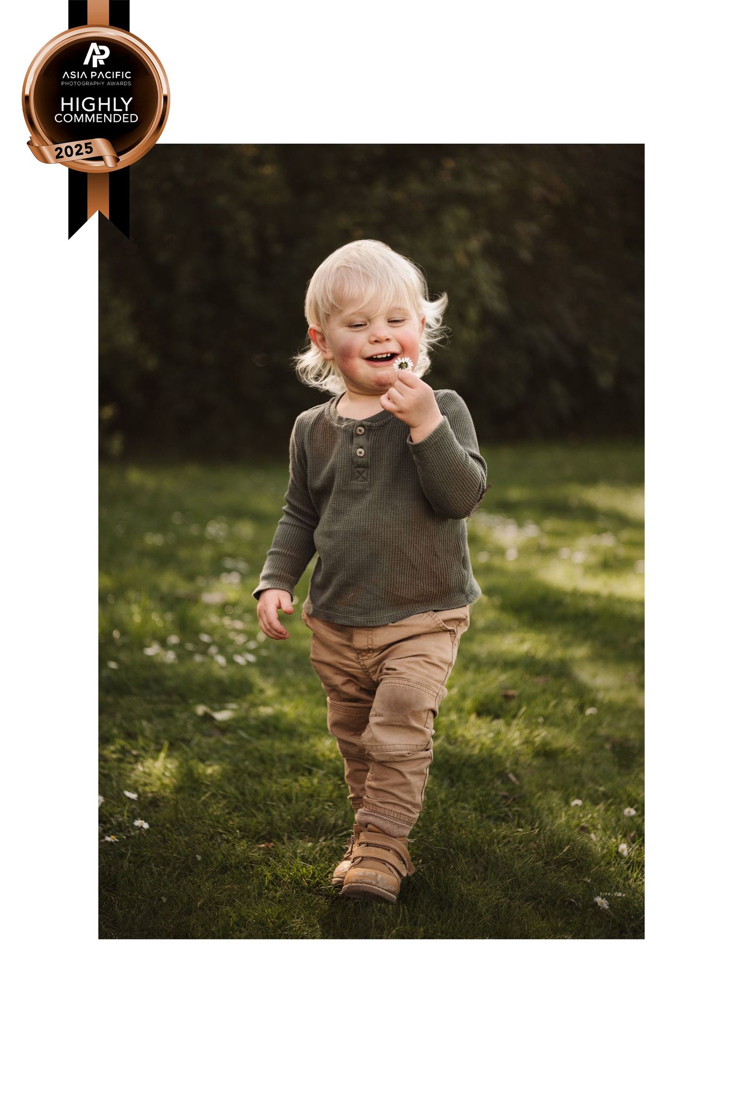 Small toddler exploring a green grass lawn while wearing brown pants and dark top on a sunny day.