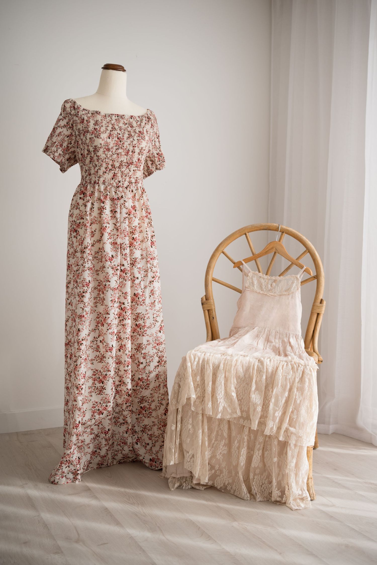 A floral maxi dress and white lace dress displayed on a mannequin and rattan chair against a white wall.