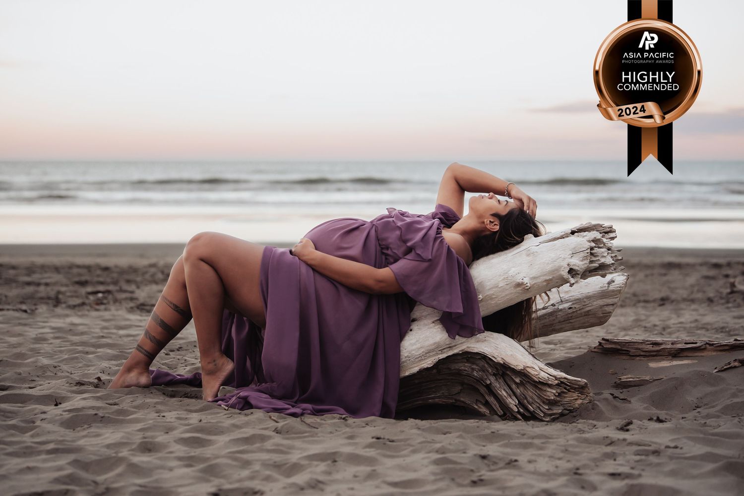Woman in flowing purple dress reclining on driftwood at a beach during golden hour.