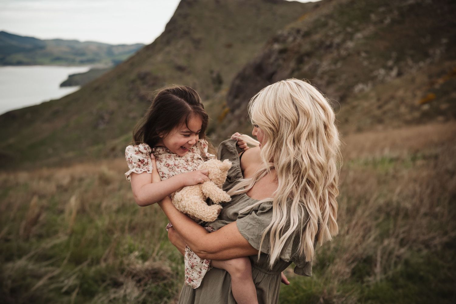 Two people share a joyful moment with a small puppy on a grassy hillside overlooking a lake.