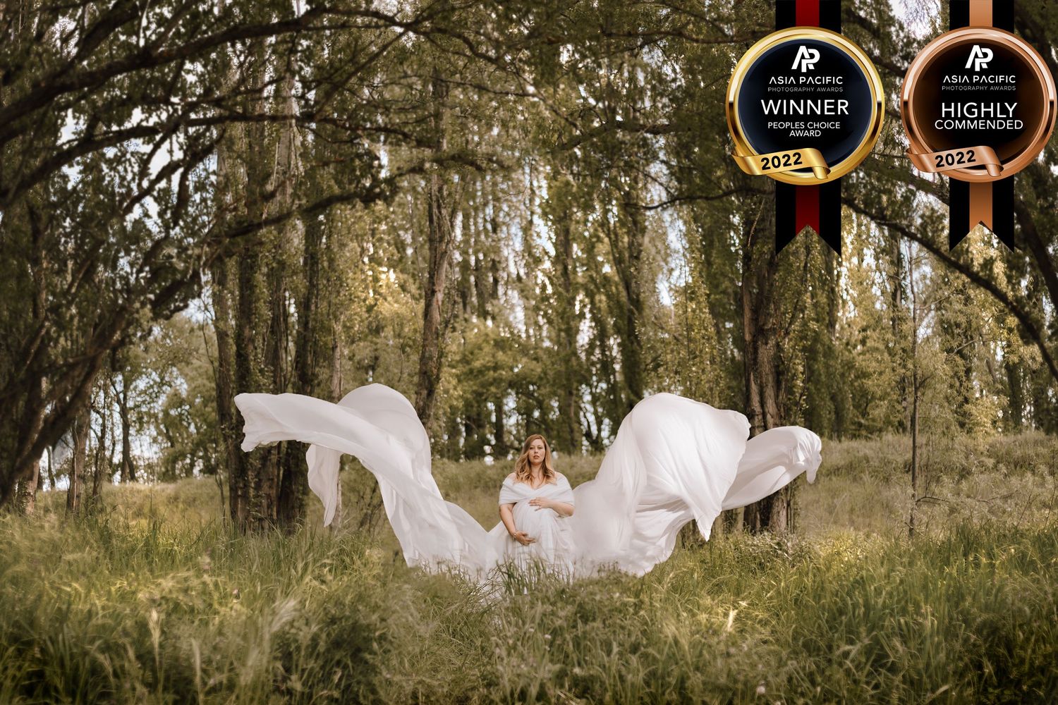 Ethereal maternity photo with flowing white fabric in wooded setting with award badges.