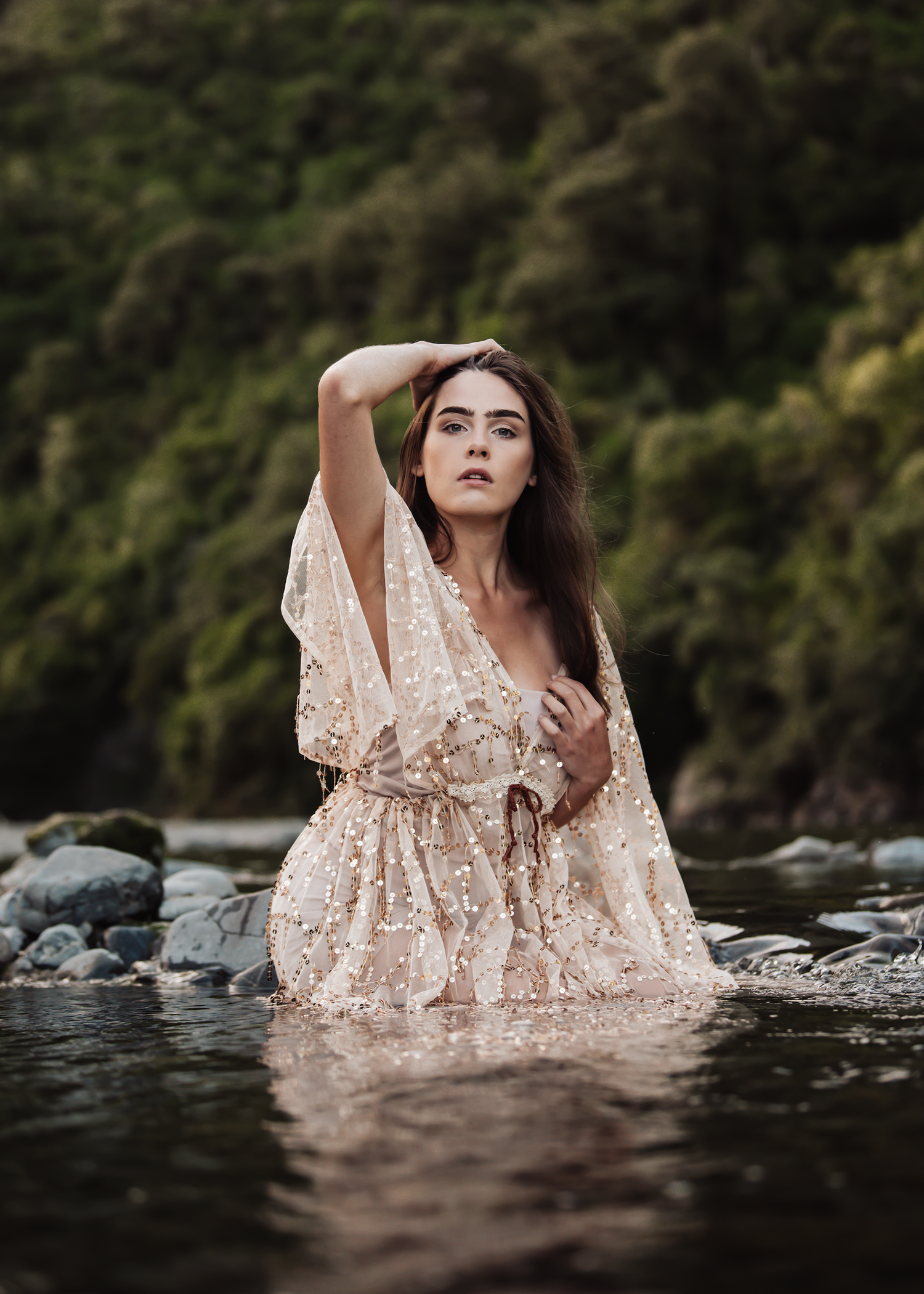 A person in a lace dress poses dramatically in a shallow river with forest backdrop.