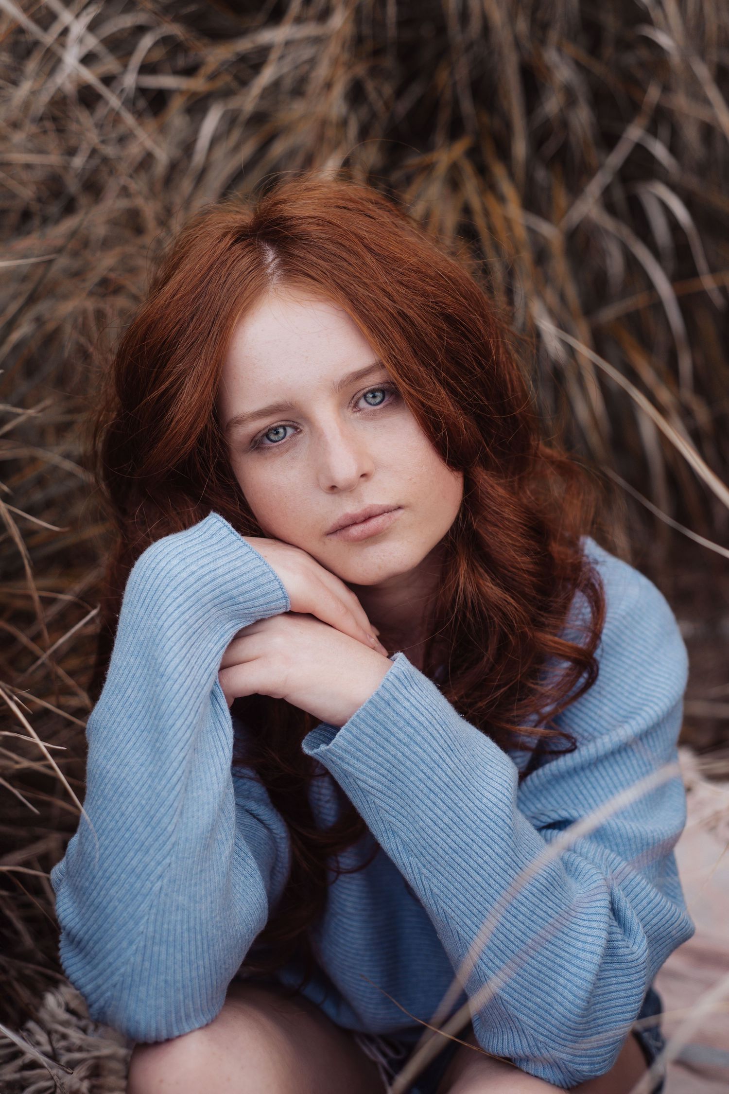Portrait of a young woman with auburn hair wearing a blue sweater against dried grass background.