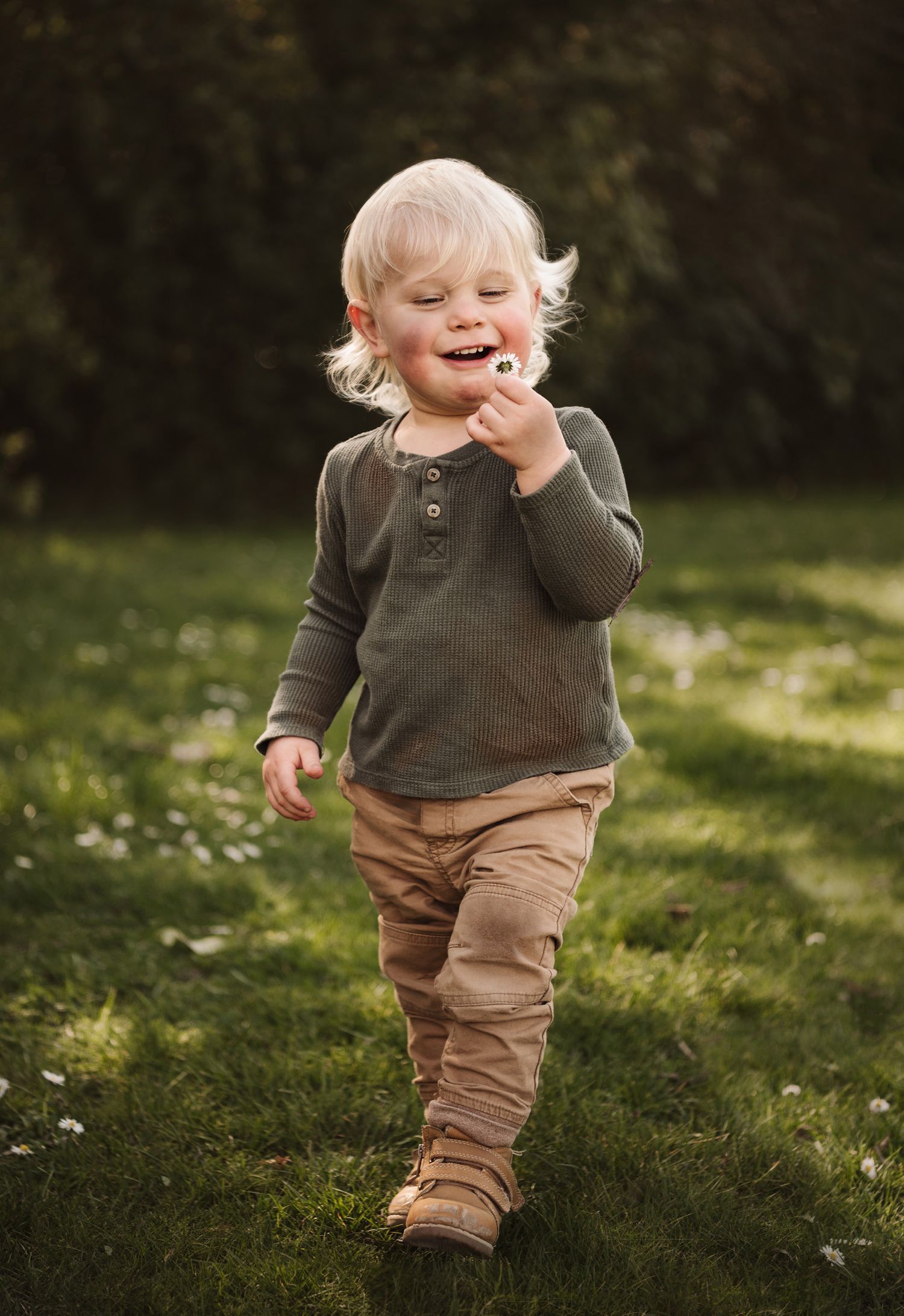 A toddler in earth-toned clothing laughs while playing outdoors in a sunny garden.