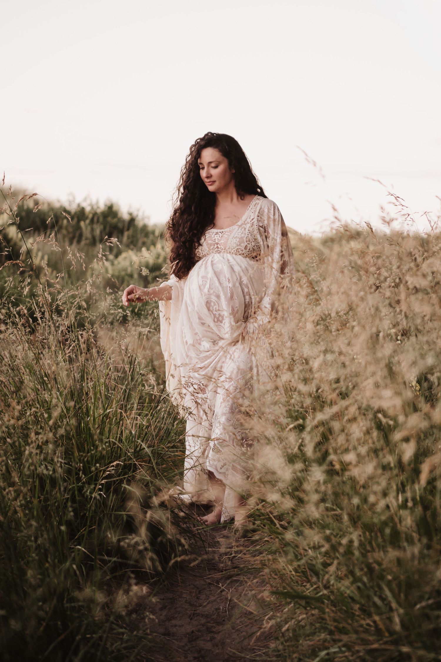 A person in a flowing white lace dress walks through a field of tall grass.