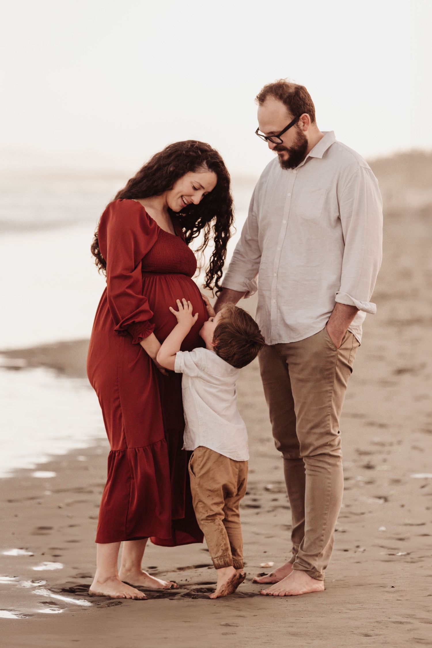 A family shares an intimate moment together on a beach at sunset, dressed in burgundy and neutral tones.