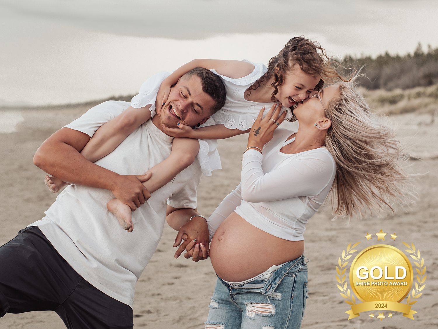 A couple in white clothing shares intimate moments on a sandy beach during a casual photoshoot.