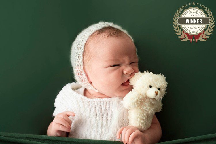 Newborn in white bonnet holding small teddy bear against green backdrop.