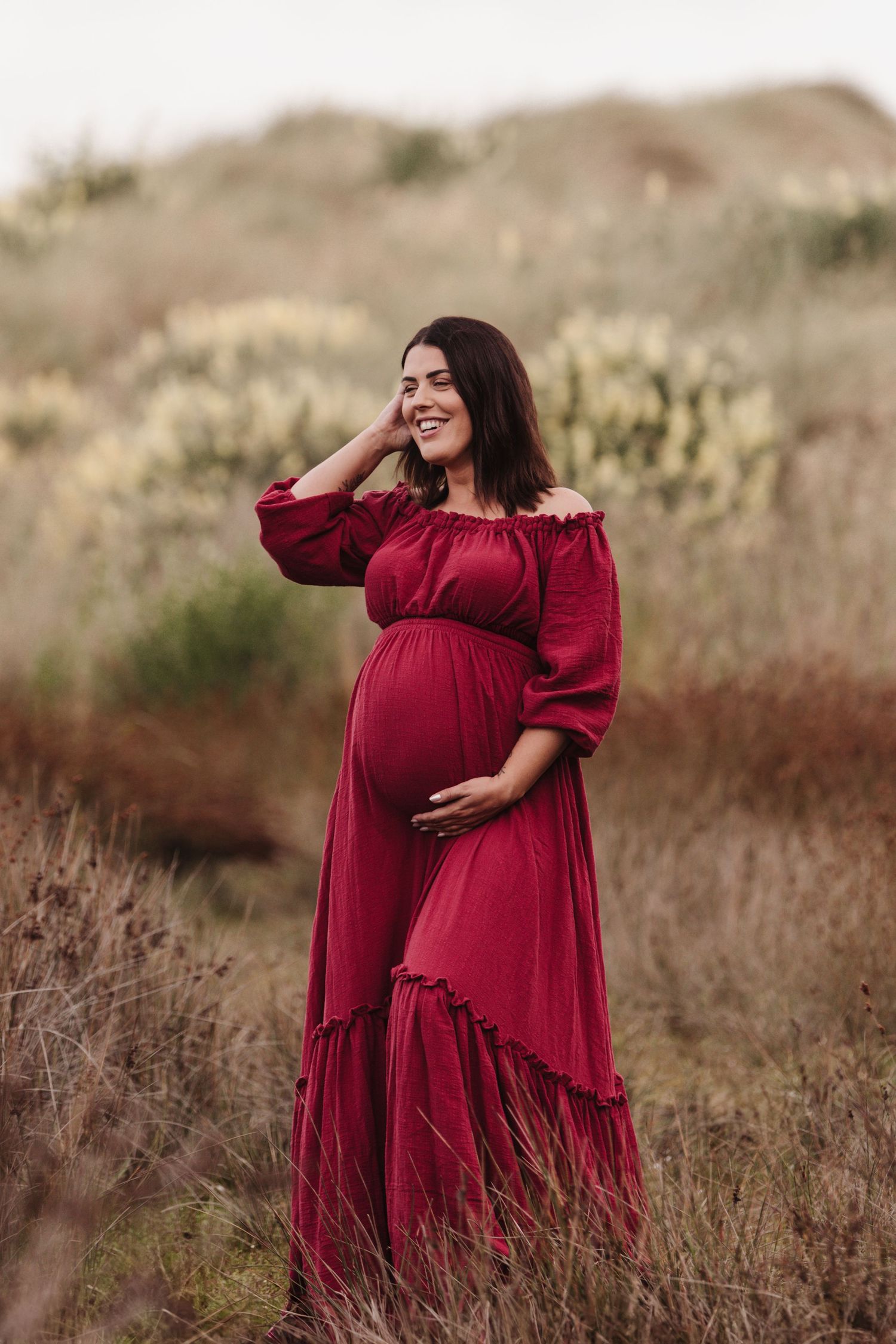 A pregnant woman in a flowing burgundy dress stands in tall dried grass during a sunset maternity photoshoot session.
