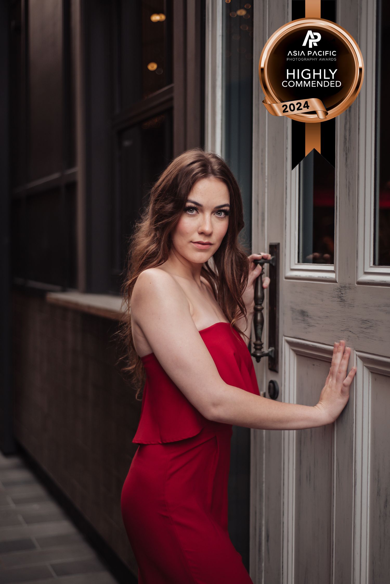 Woman in elegant red dress posing against a grey wall in dramatic lighting.
