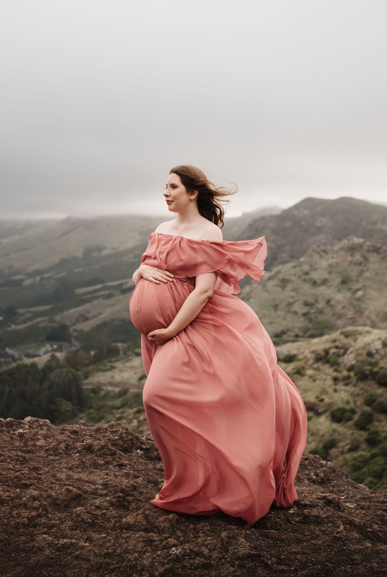 A person in a flowing pink gown stands gracefully on a rocky mountain peak with dramatic cloudy skies in the background.
