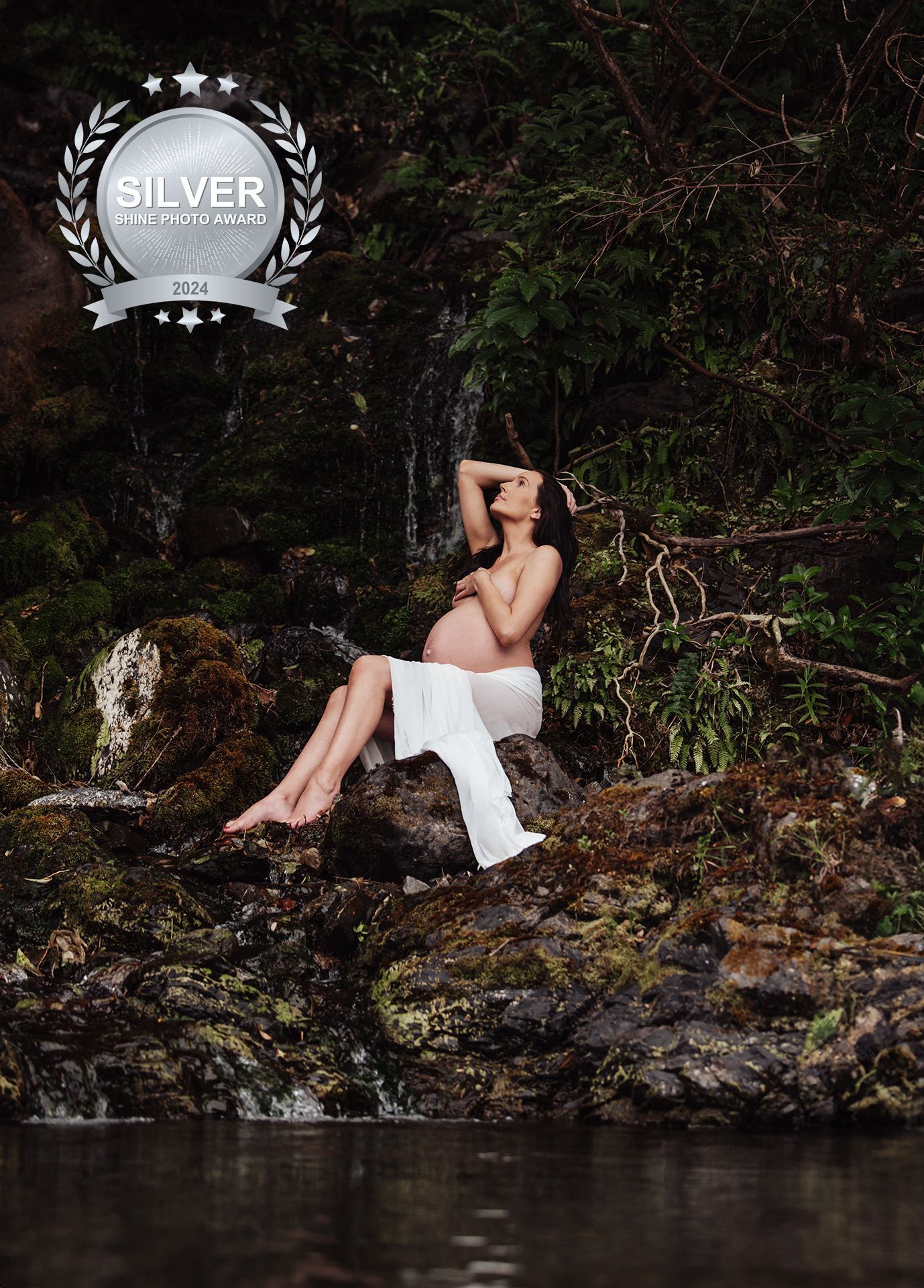 A person in white clothing reclines on mossy rocks along a dark riverbank at twilight.