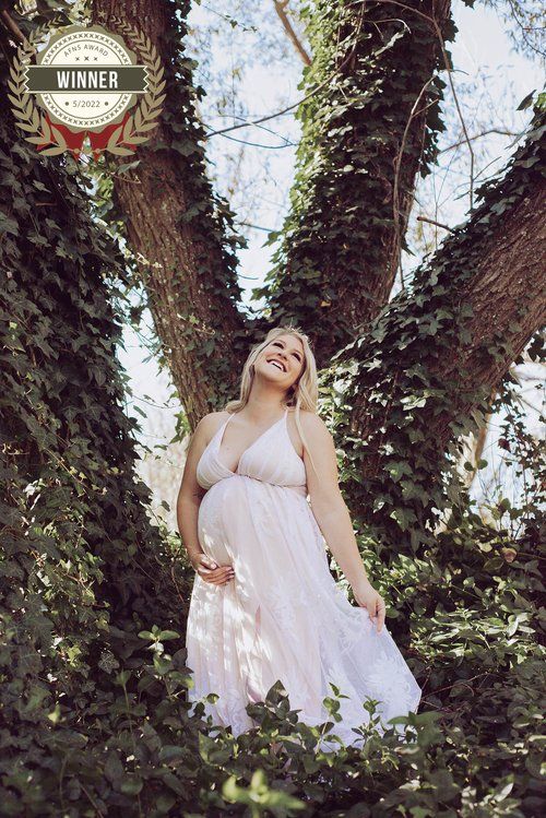 Maternity portrait of woman in white dress standing between ivy-covered trees.