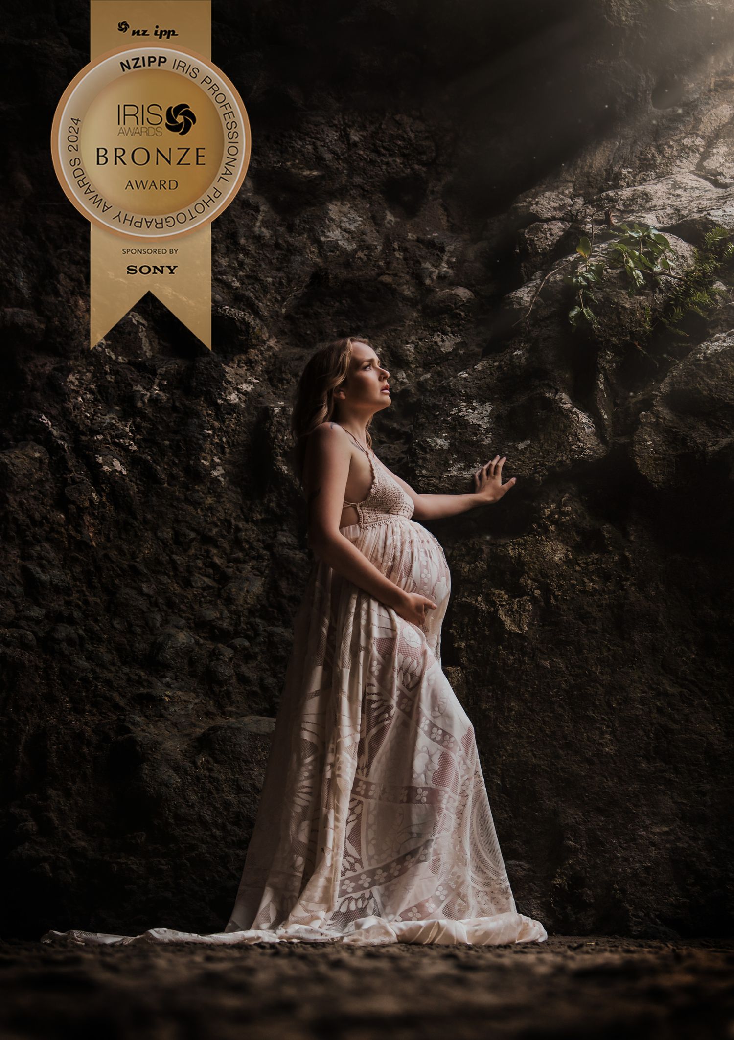 Ethereal maternity portrait of woman in white lace gown against dramatic rocky backdrop.