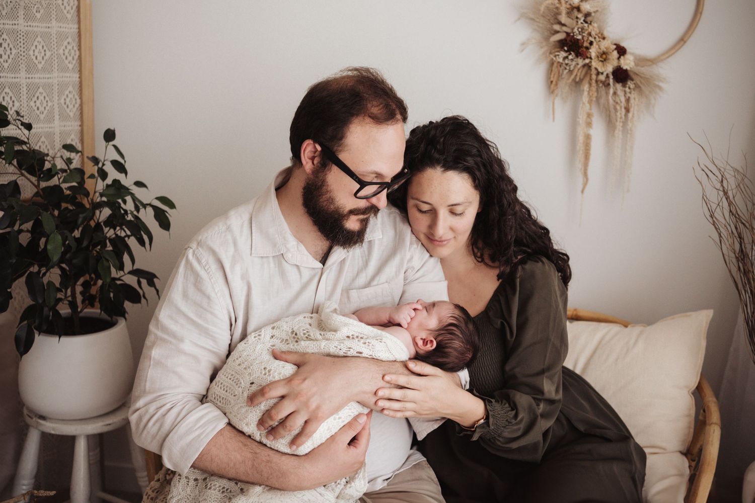 A couple in casual attire holds and gazes lovingly at their newborn baby while sitting together on a couch.