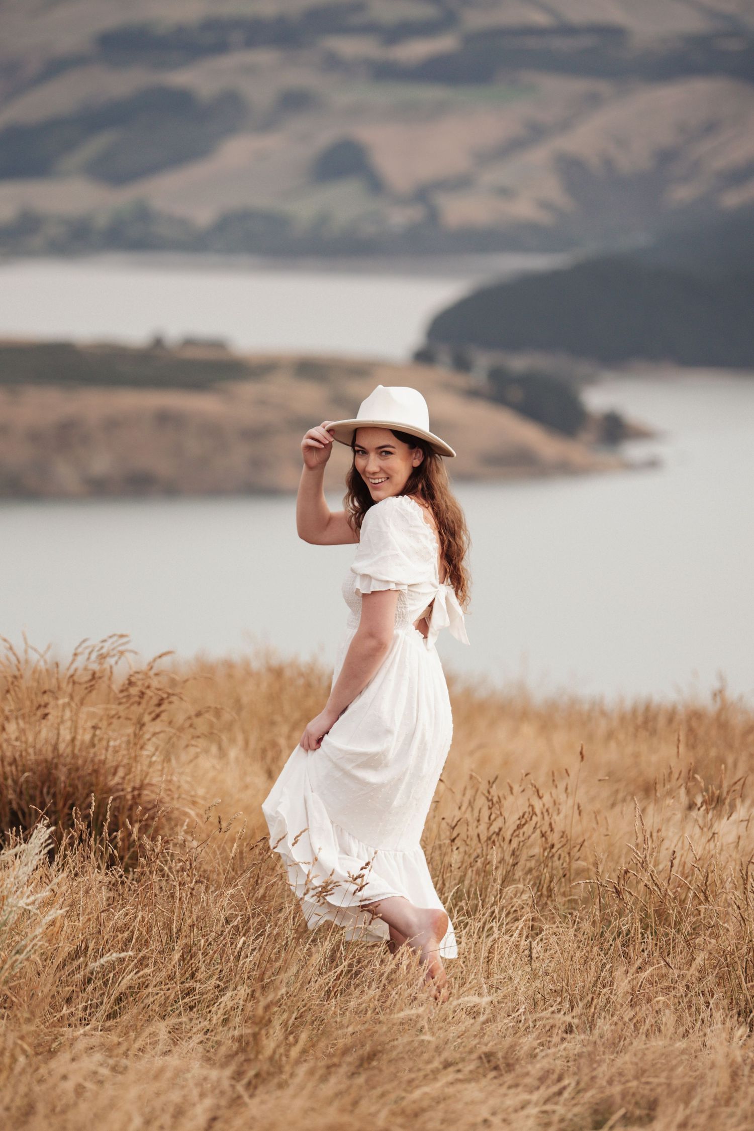 Woman in white dress twirls on hilltop with water view behind her.