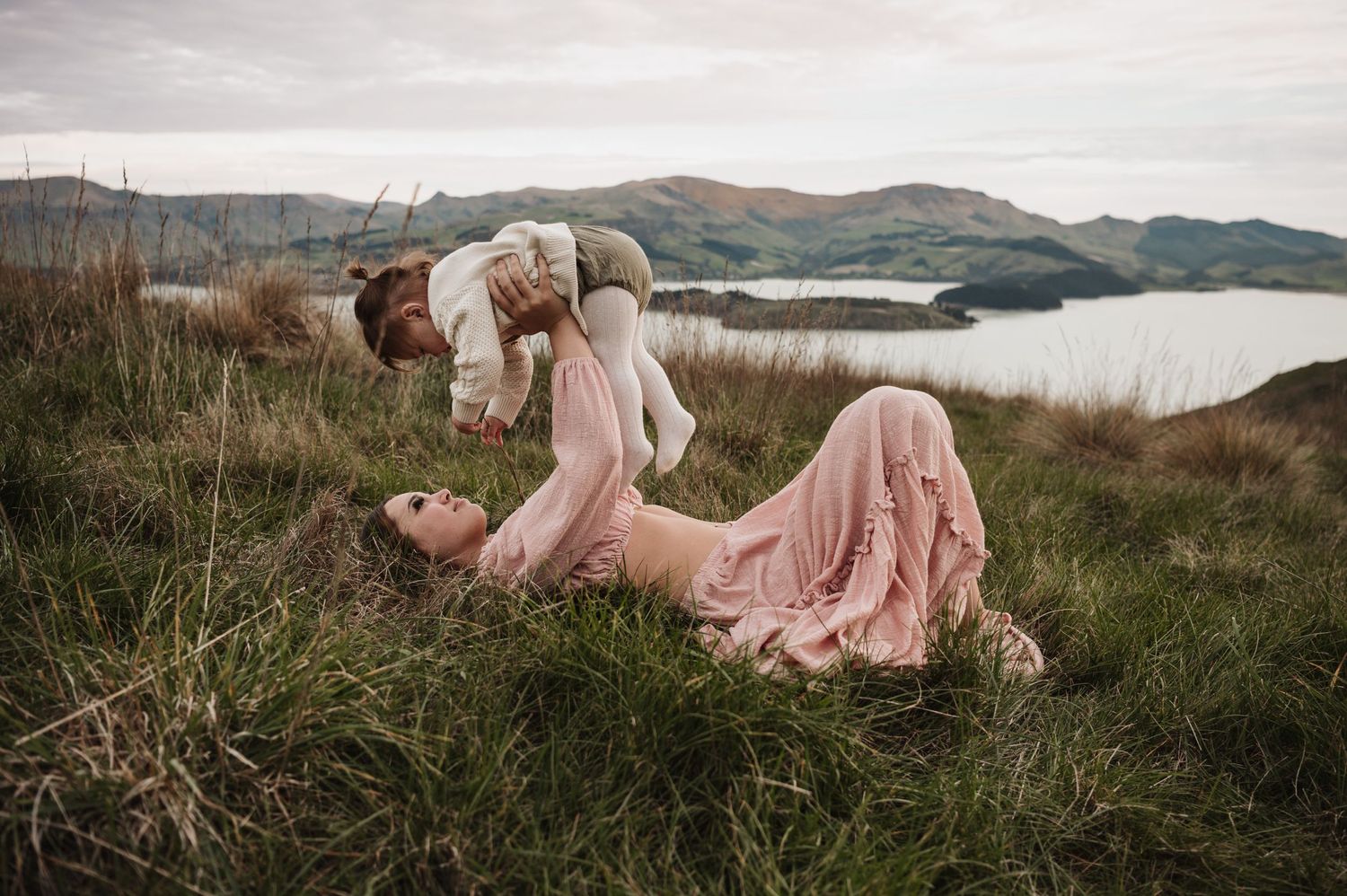 Person in pink dress lies in grass on scenic hilltop overlooking mountains and lake at dusk.