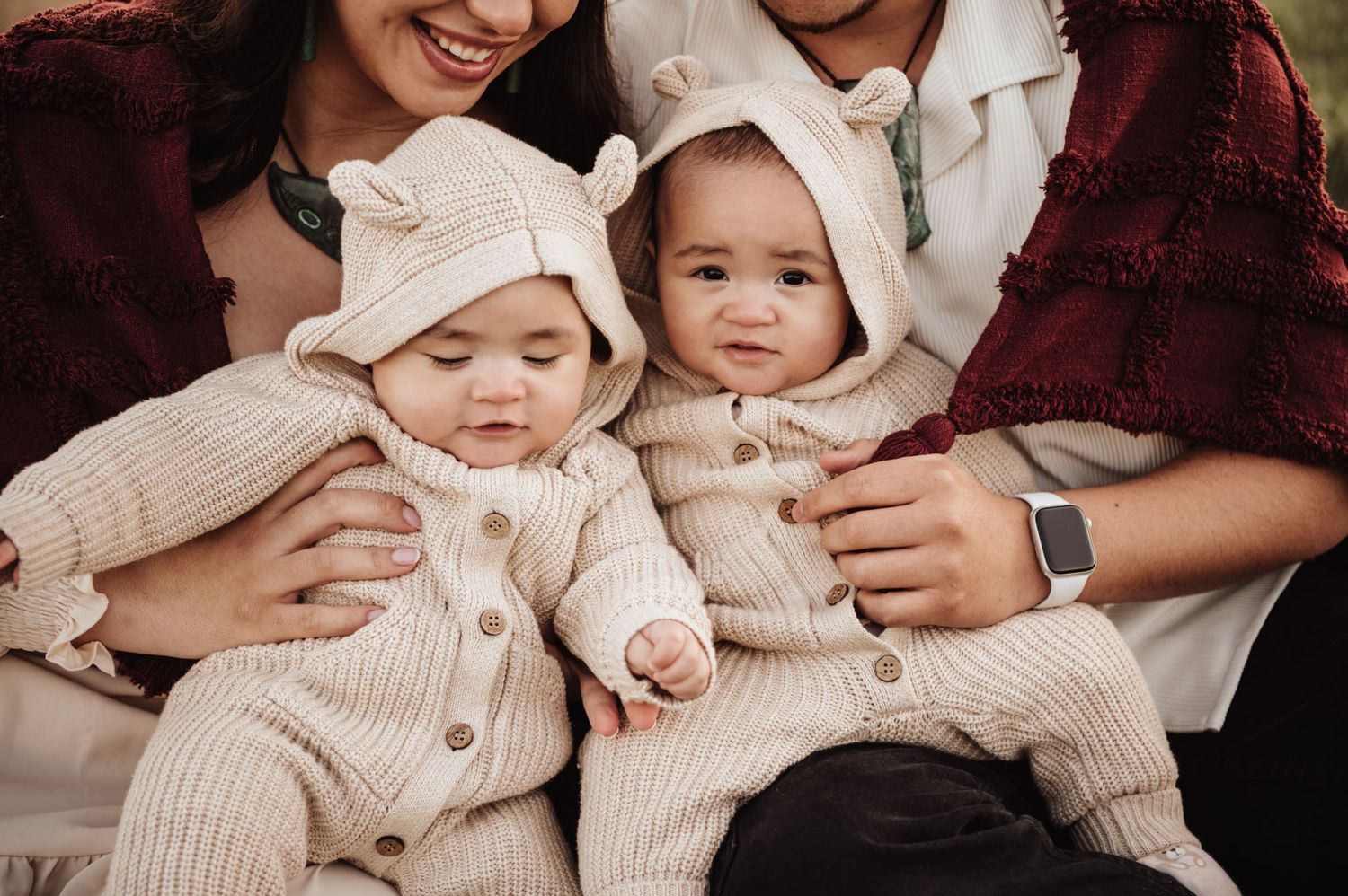 Two babies wearing matching cream knit outfits and bonnets sit together with family members under a burgundy blanket.