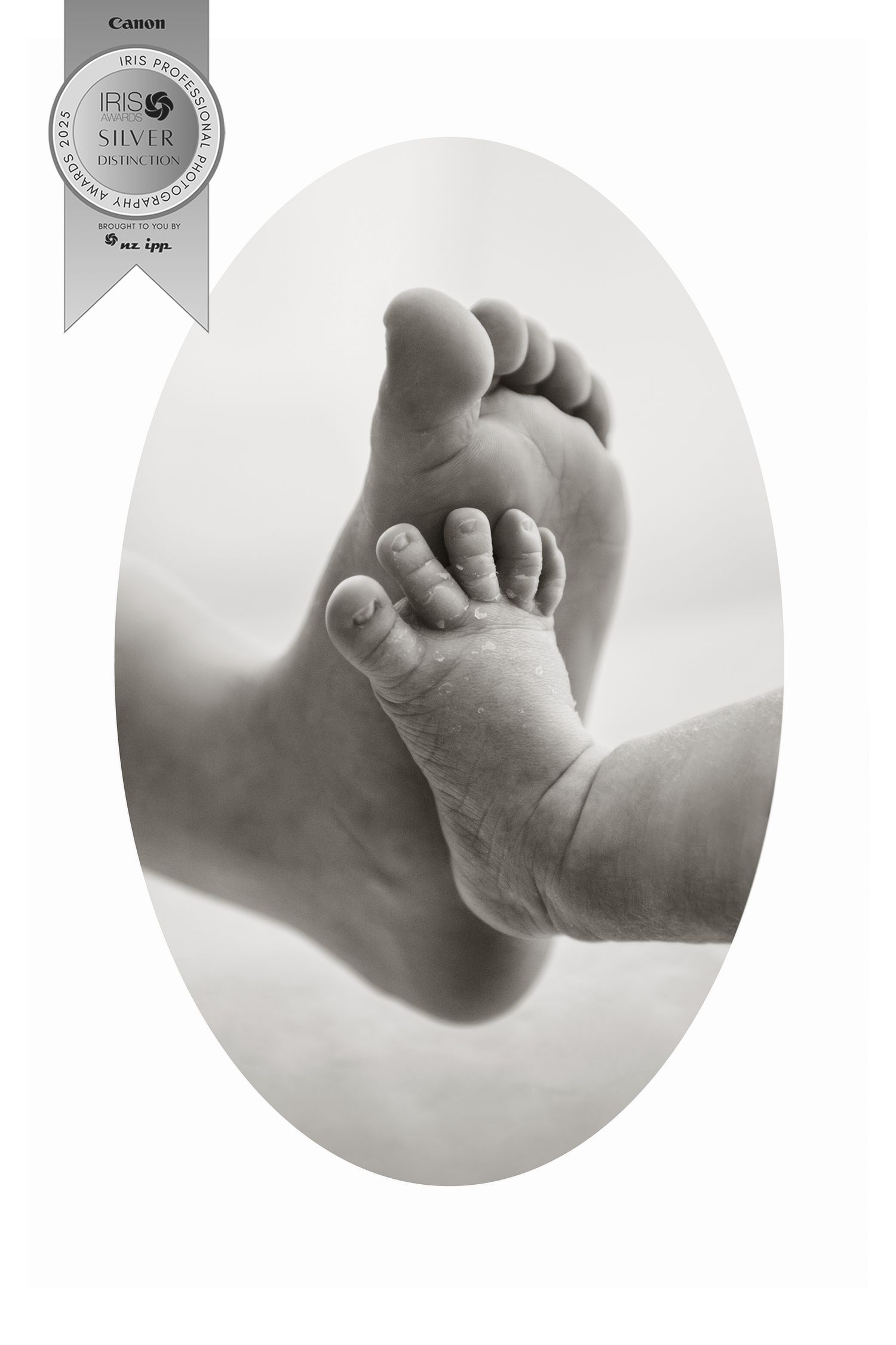 A black and white artistic photograph showing two touching feet, one adult and one baby foot, in an oval vignette.