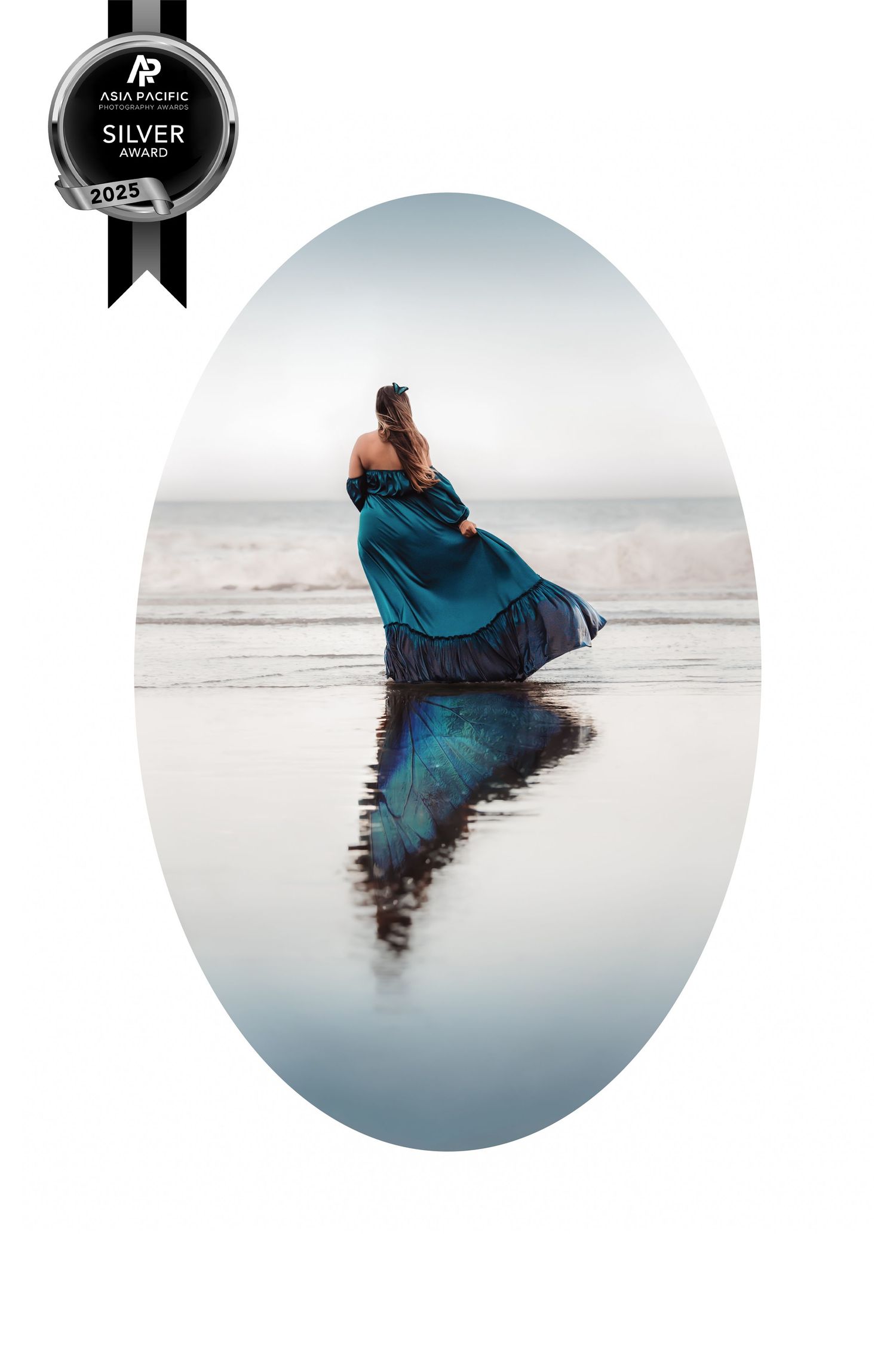 A figure in a flowing blue dress stands on a reflective beach at sunset creating a mirror-like reflection on the wet sand.