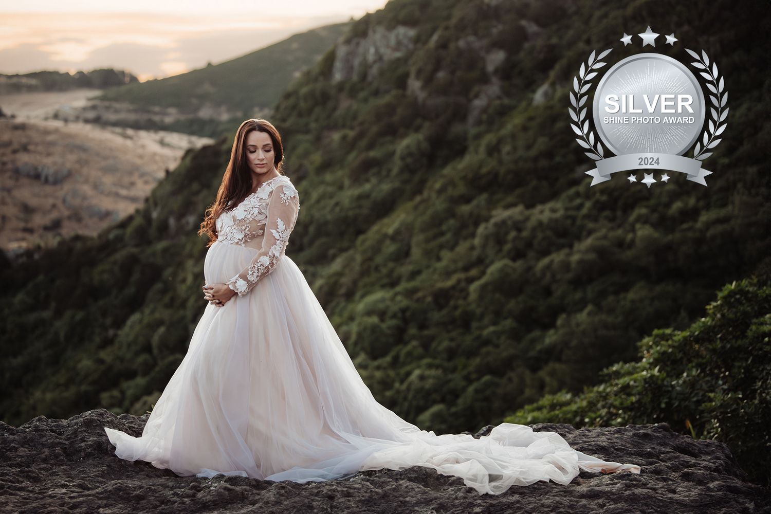 Elegant maternity photo of woman in flowing white gown standing on mountain cliff at sunset.