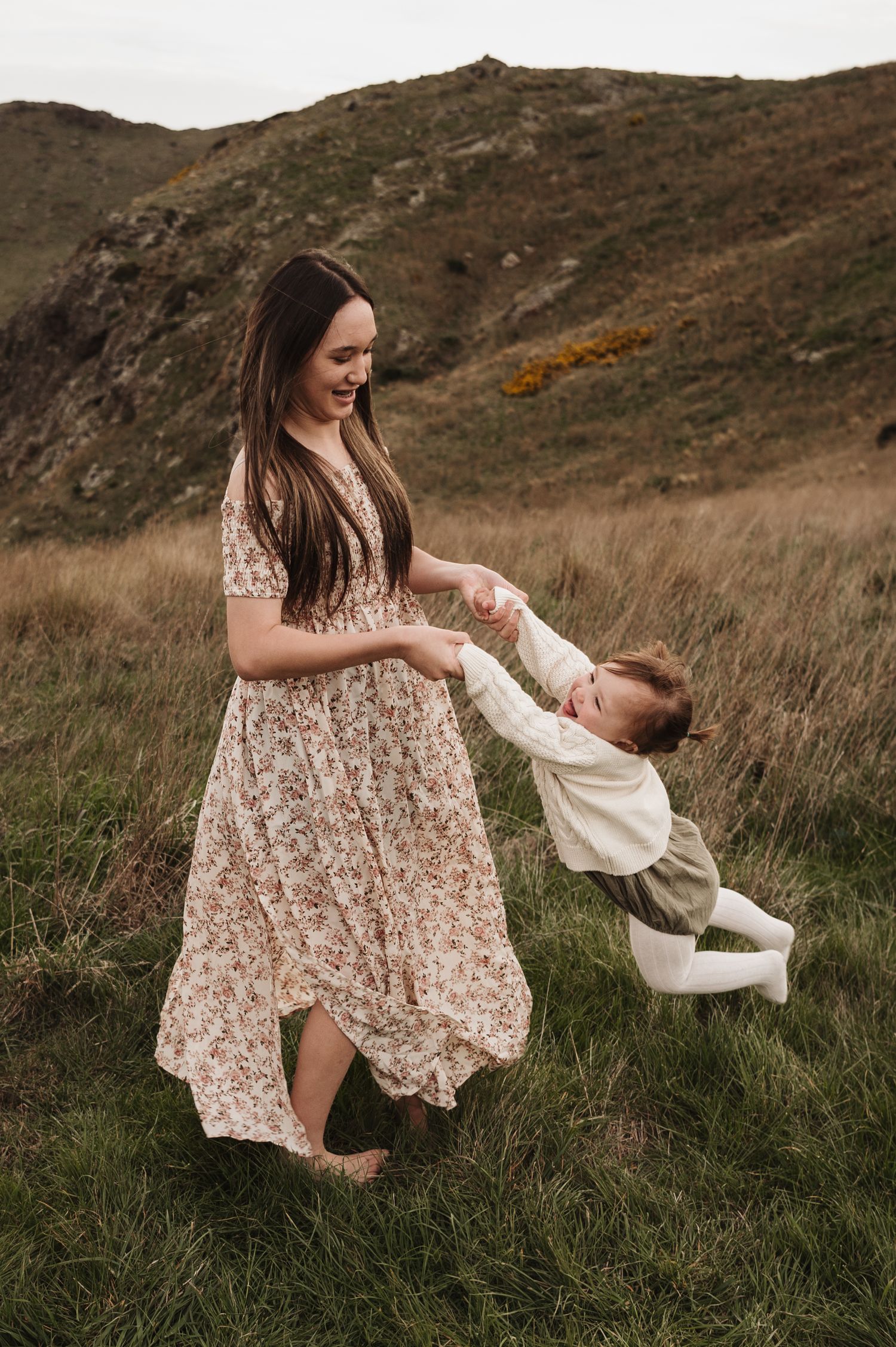 A woman in a floral dress dances with a toddler in white clothing on a grassy hillside at sunset.