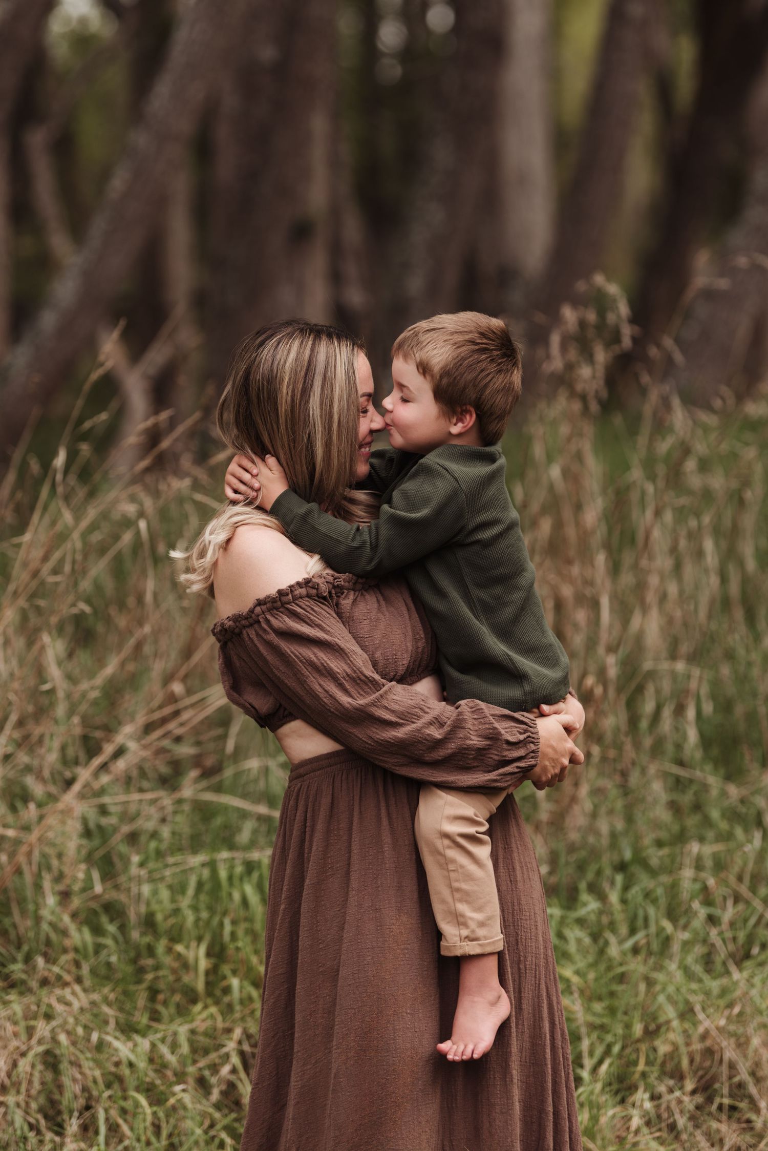 A tender embrace shared between two figures in a brown dress and green sweater among tall grass and trees in the forest.