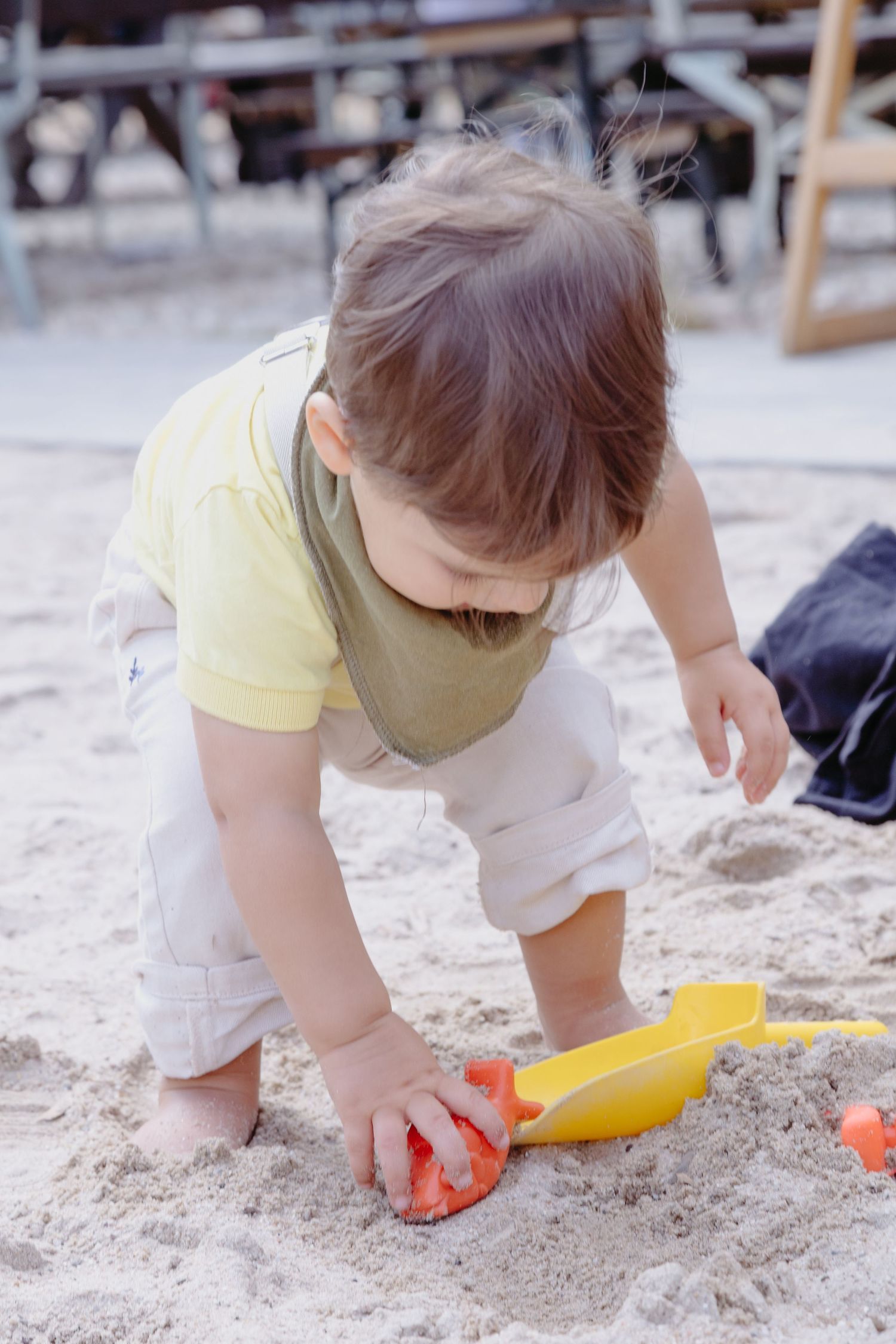 A small child in a yellow shirt and white shorts plays with colorful toys in a sandbox.
