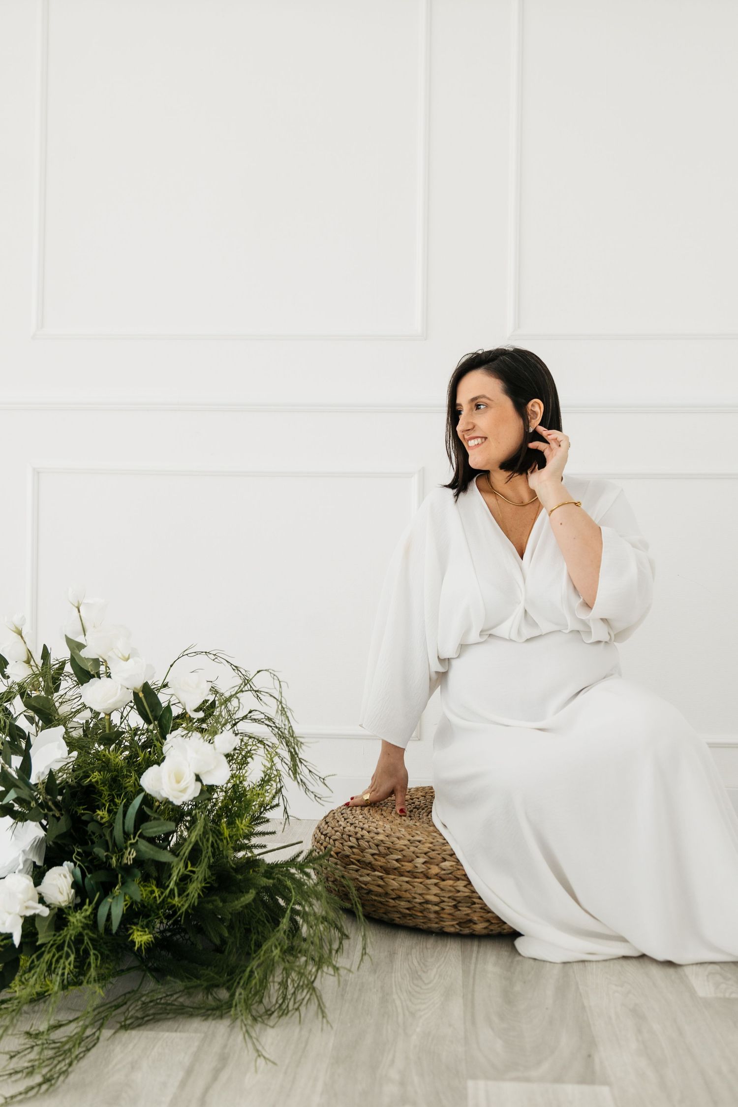 A figure in white loungewear rests on a light wooden floor beside greenery and a woven basket in a bright setting.
