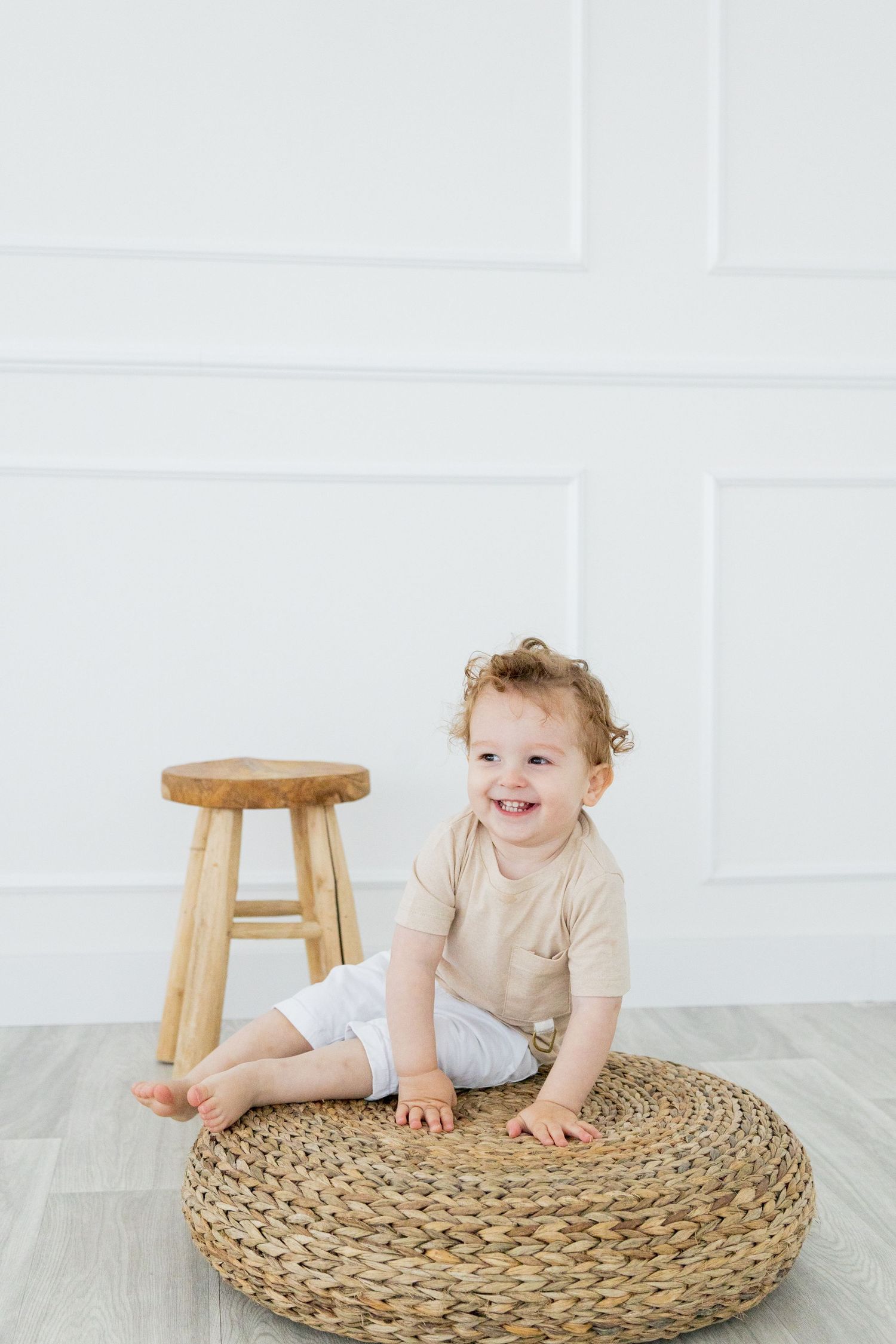 Small child sitting on a round woven ottoman beside wooden stool in minimalist white room setting.